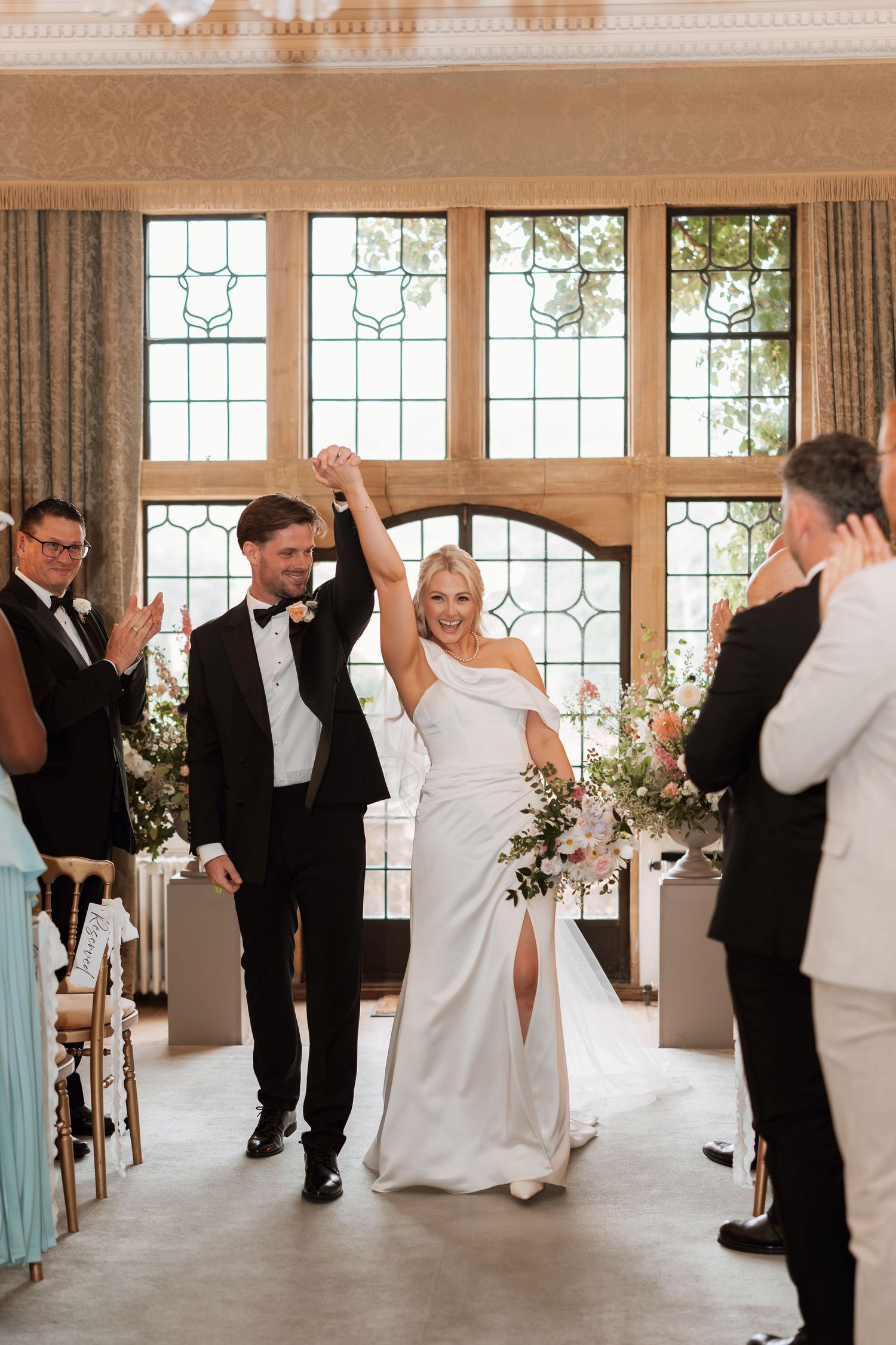 Newlyweds celebrate with raised arms while exiting down the aisle in an elegant venue with stained glass windows.