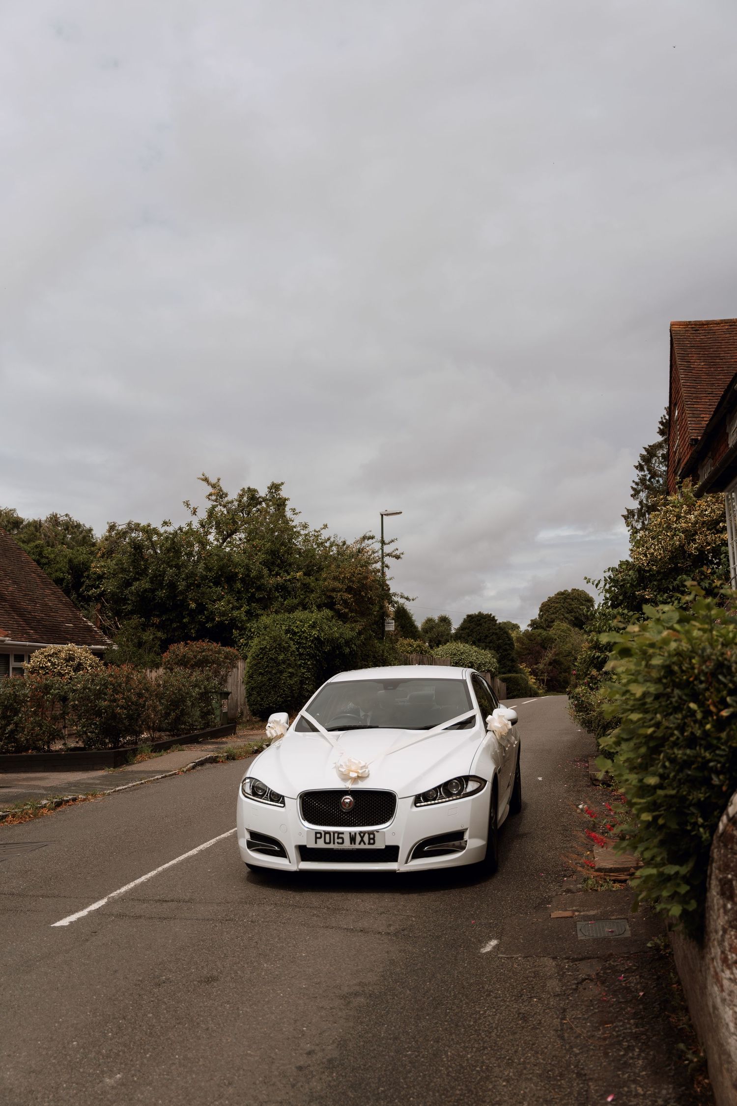 A white Jaguar luxury sedan parked on a residential street with houses and greenery visible in the background.