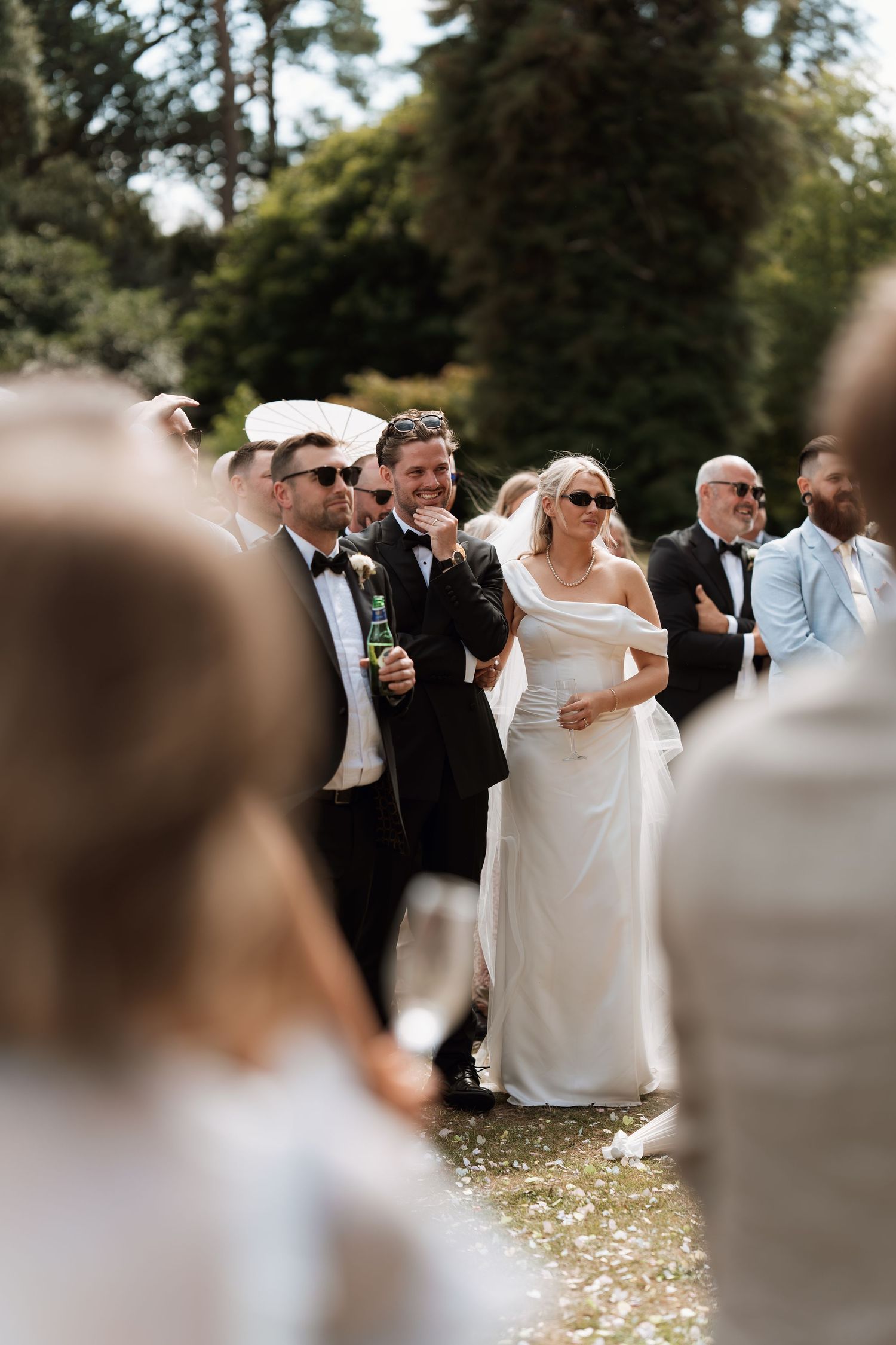 Wedding ceremony guests gather outdoors under tall trees on a sunny afternoon.