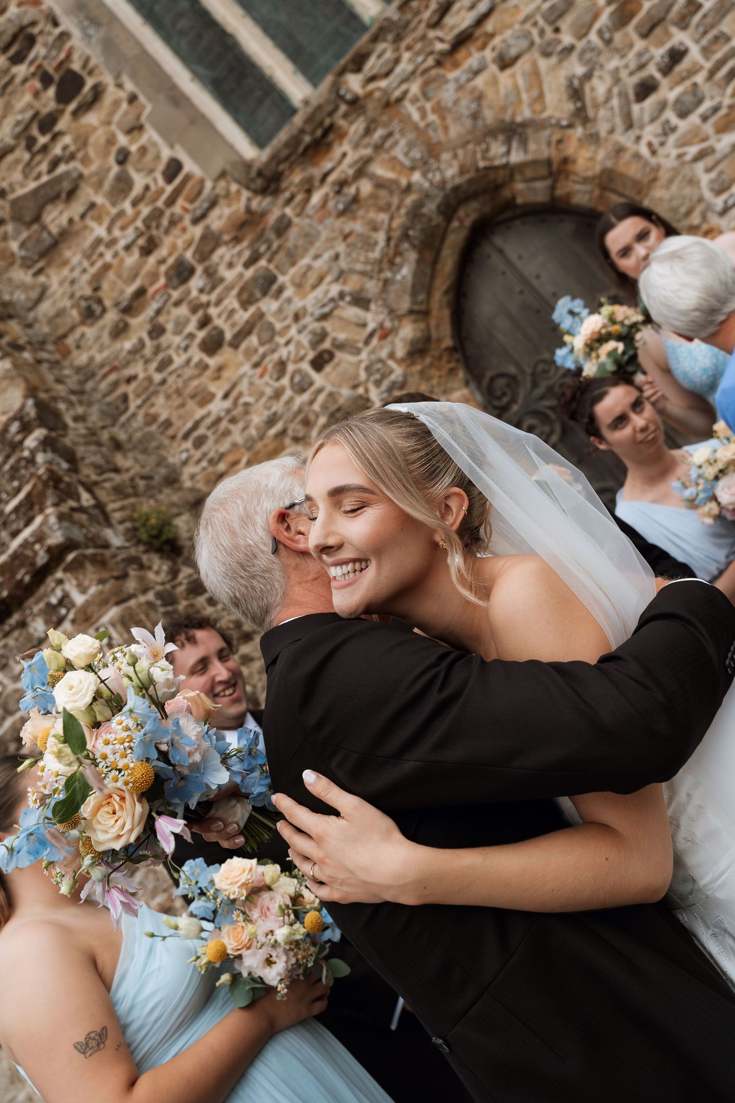 An emotional wedding moment captured against a rustic stone wall with delicate blue and cream floral bouquet details.