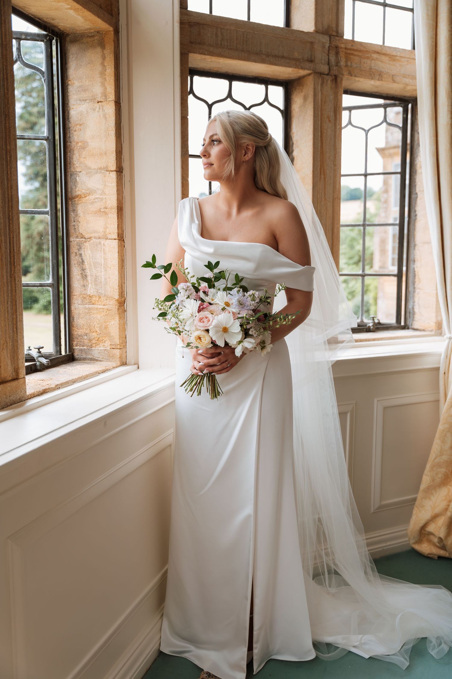 Elegant bride in off-shoulder white gown holds lush floral bouquet while standing by ornate vintage windows in stone interior.