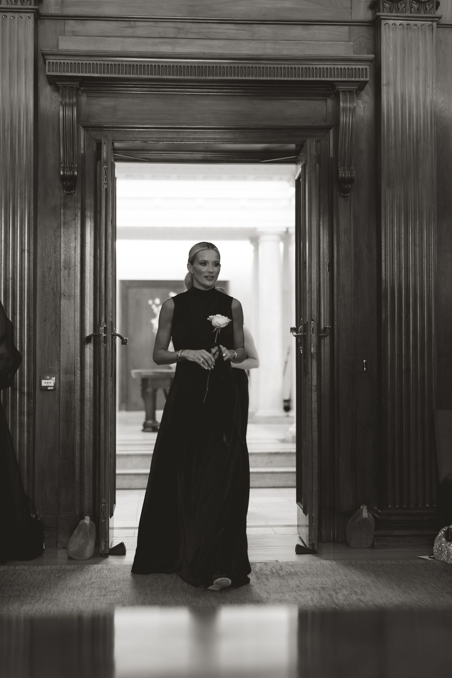 Artistic black and white photograph showing figure walking through elegant hallway with wooden paneling.
