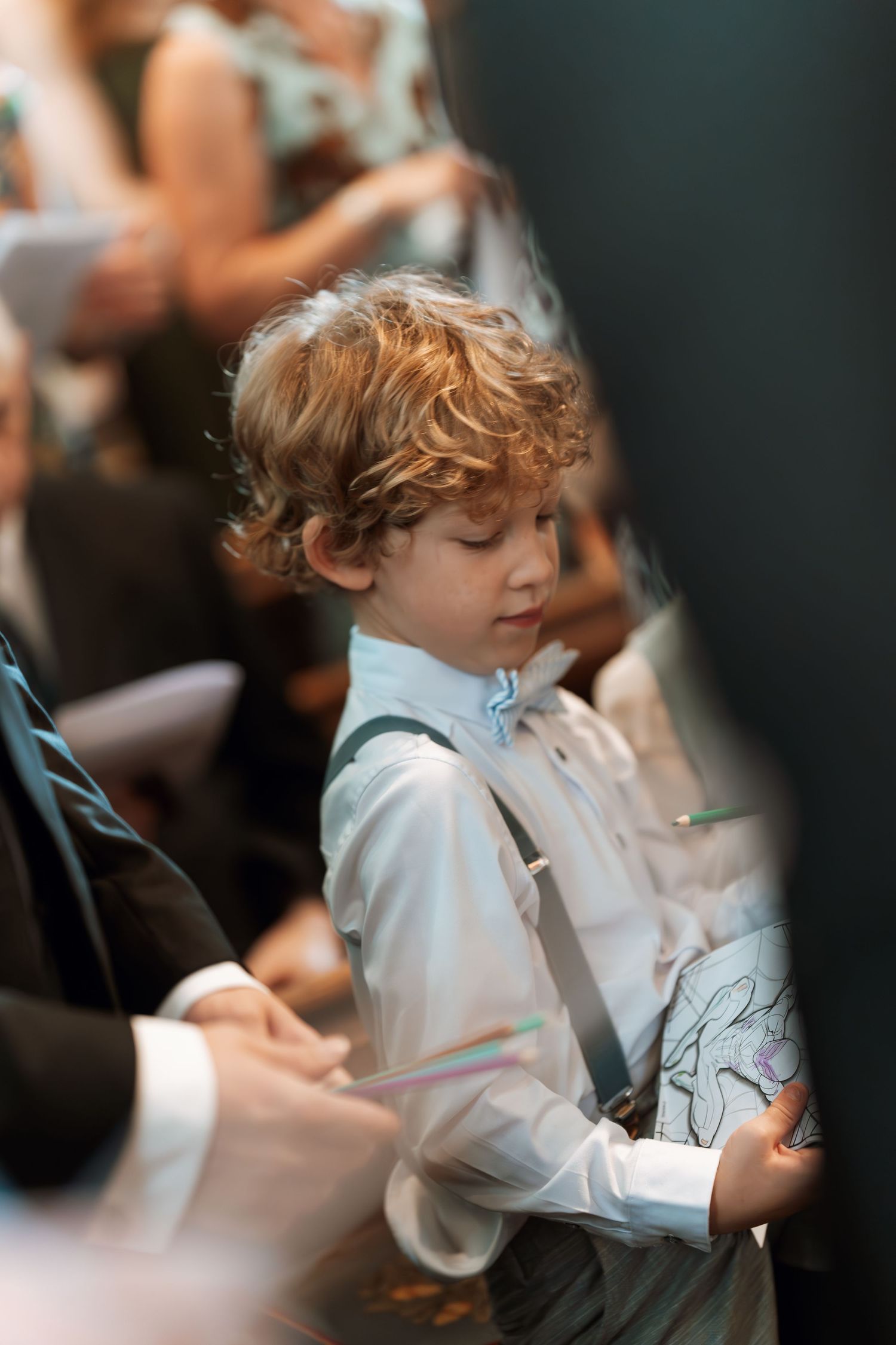 Young boy in white dress shirt and suspenders at formal event looking down thoughtfully.