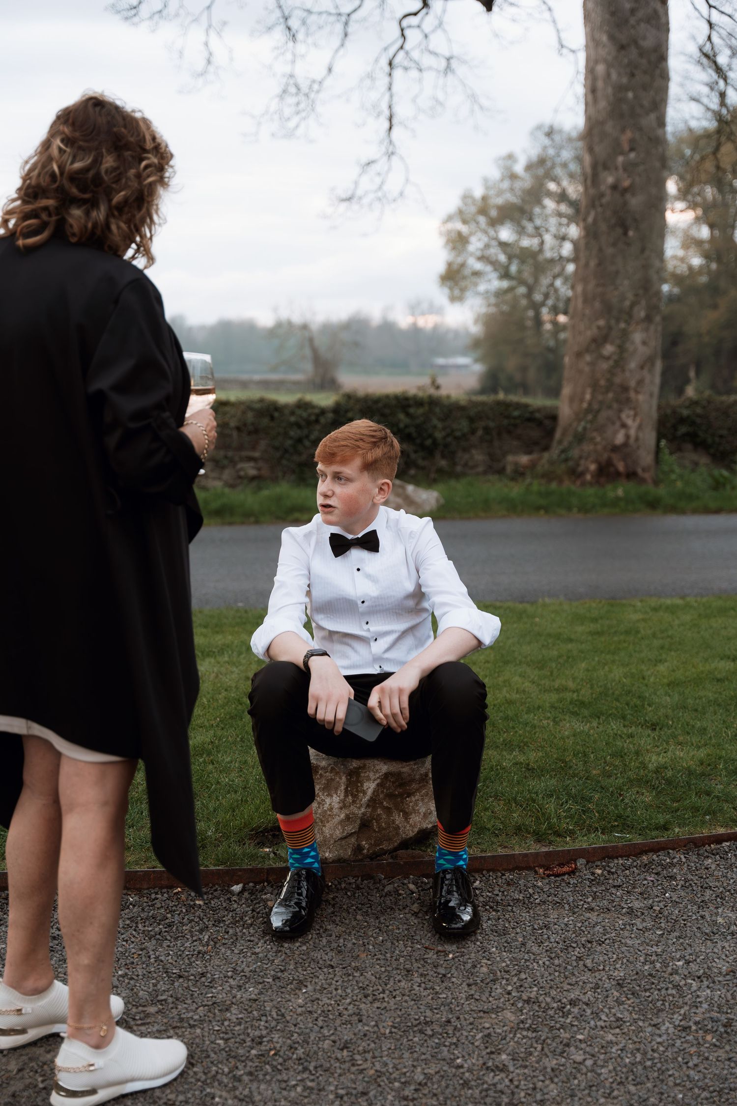 Young guest in formal wear sits on tree stump during outdoor event.
