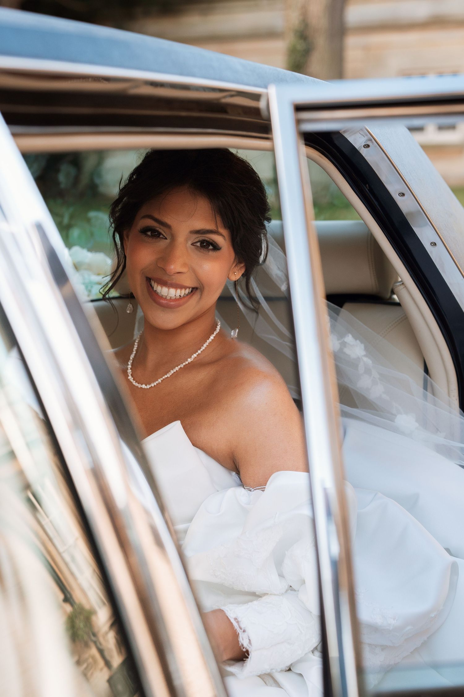 Woman in white wedding dress and pearl necklace sitting in classic car.
