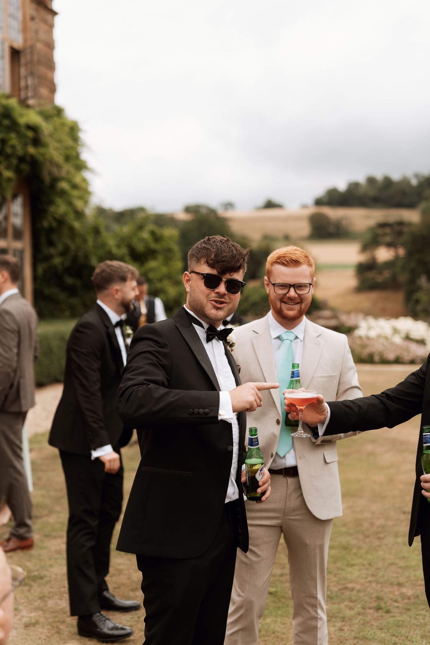 A group of groomsmen in suits and tuxedos gather outdoors on a sunny day in front of rolling hillsides.