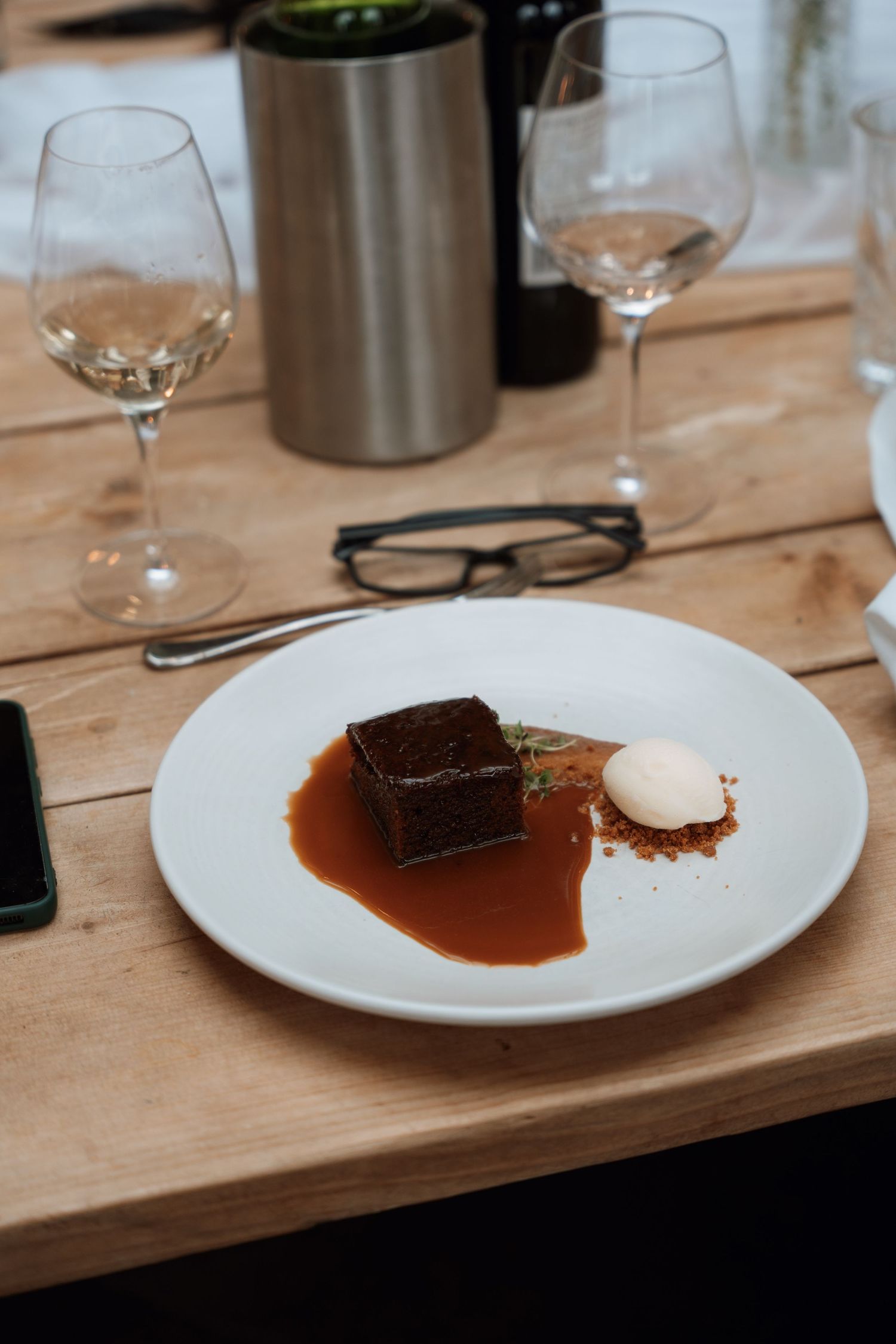 Chocolate dessert with caramel sauce and cream served on white plate with wine glasses and reading glasses.