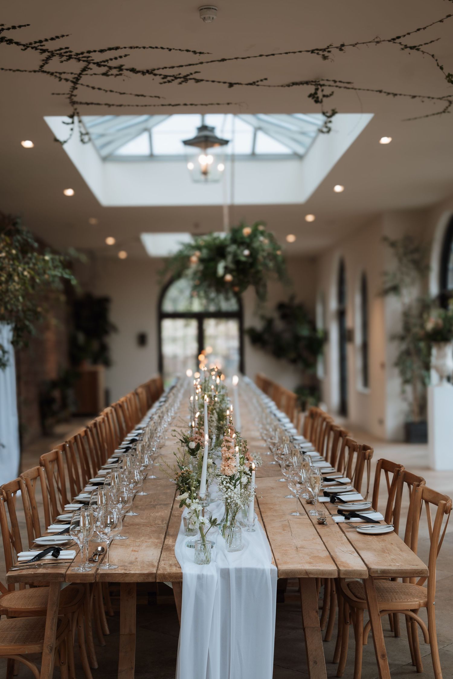Elegant wedding reception table setup with wooden chairs and white runner under skylight.