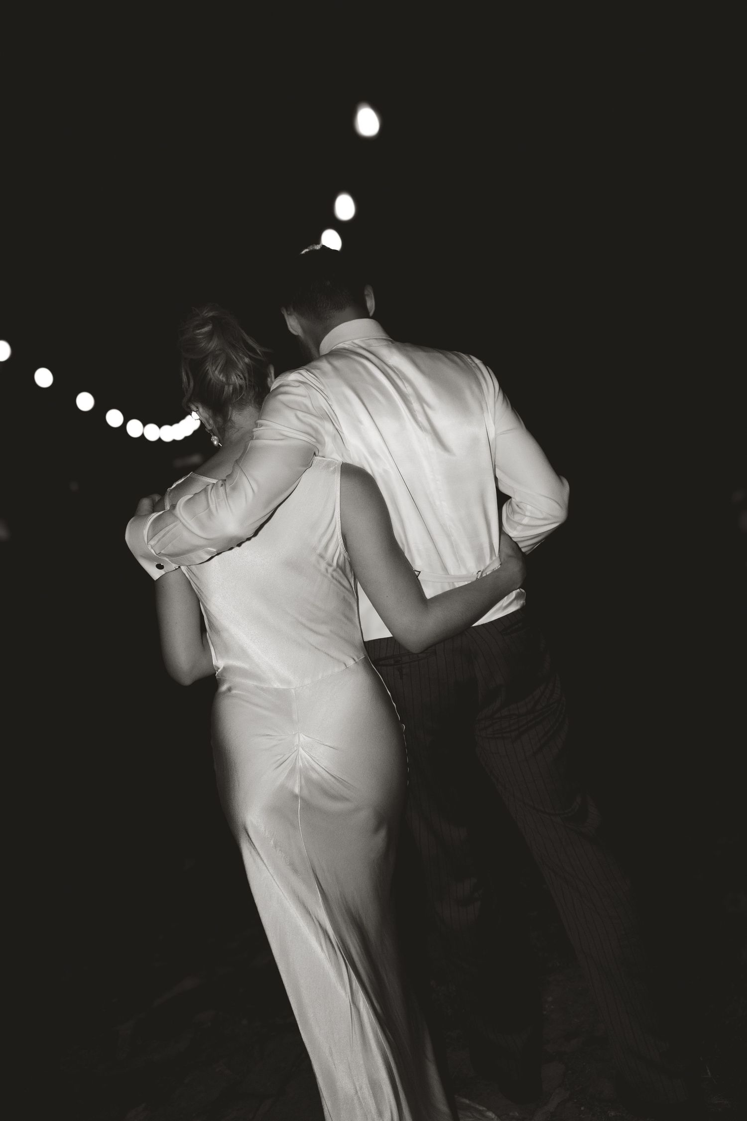 A black and white photo of a couple dancing under string lights at their wedding reception.