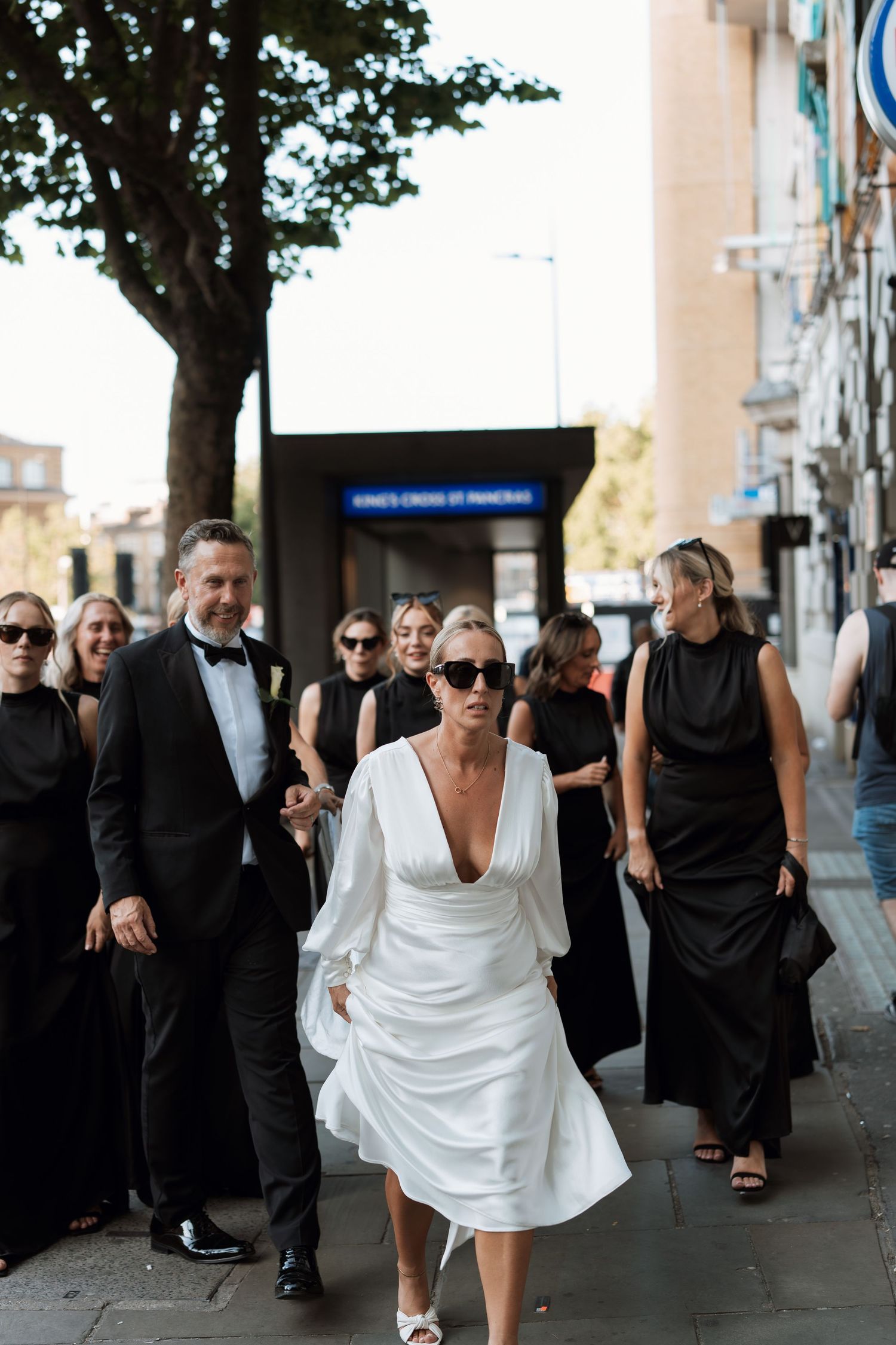 Wedding party in formal attire walking down a city street with the bride in a flowing white dress.