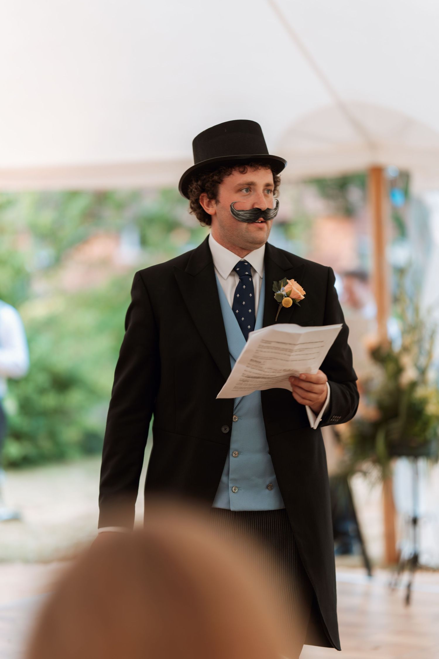 A speaker in vintage attire with top hat and fake mustache reads from a paper during the wedding celebration.