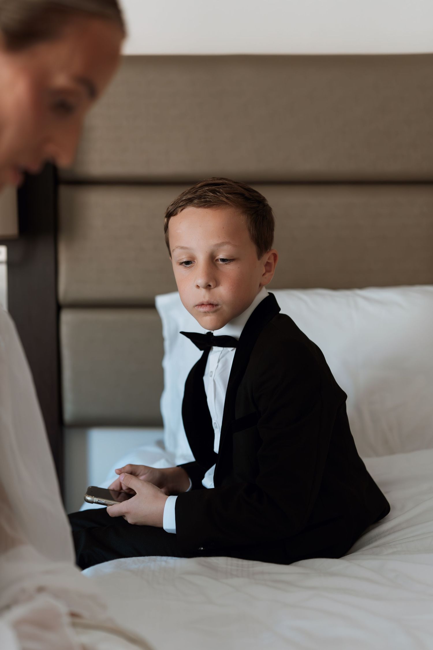 Young person in formal black attire resting on white hotel bedding while looking at mobile device.
