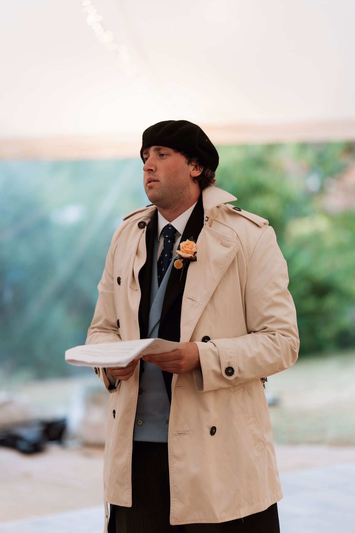 A person in a beige trench coat and winter hat holds papers during an outdoor event.