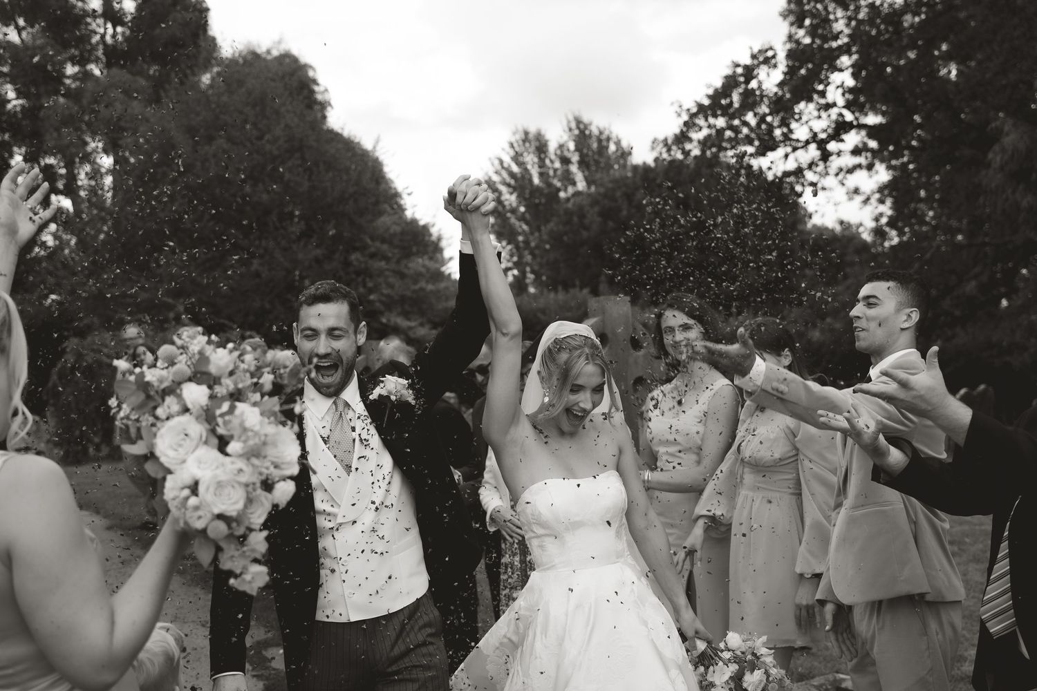 Joyful newlyweds and wedding party celebrate with outstretched arms during outdoor garden celebration.