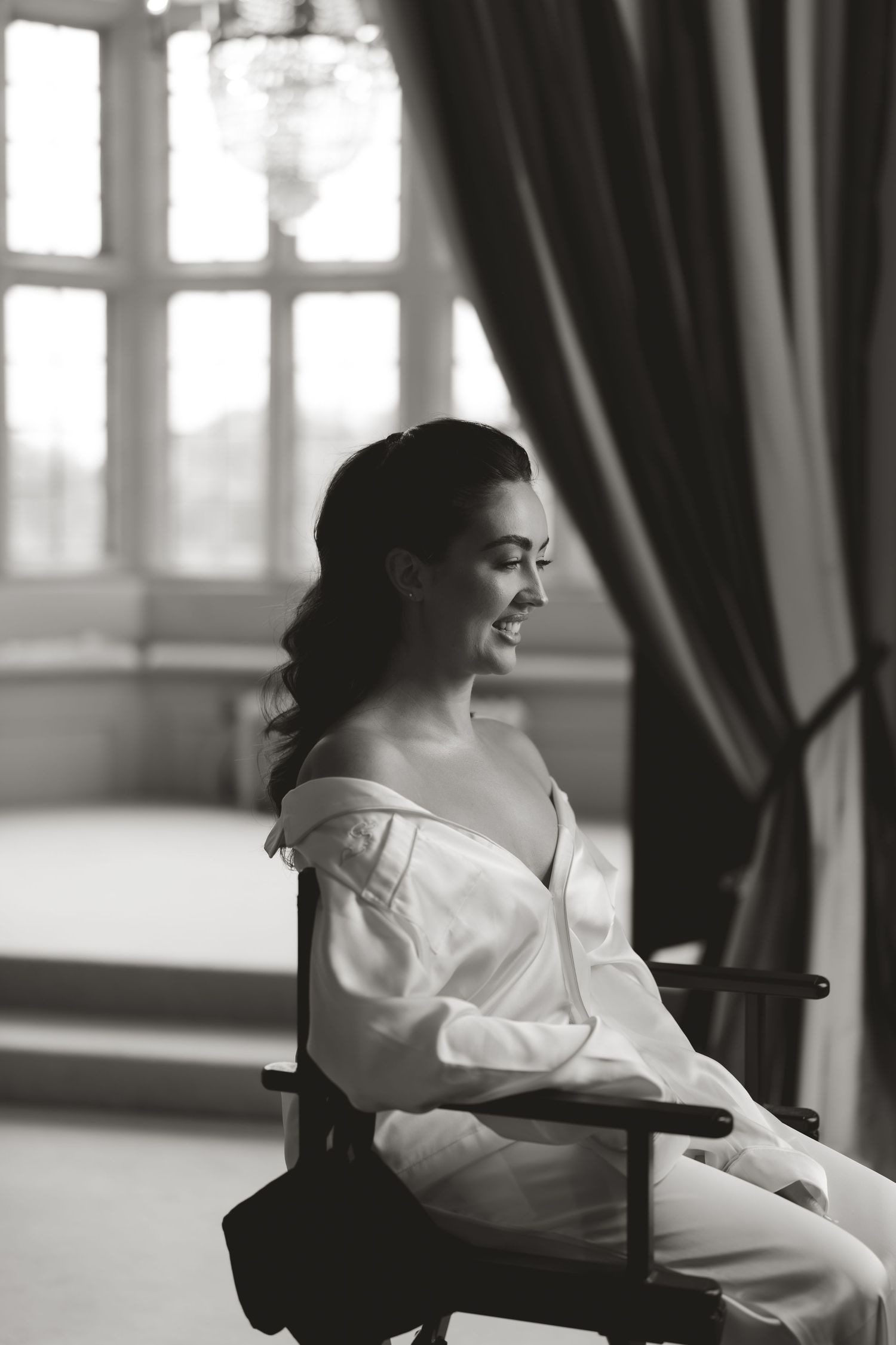 A black and white portrait of a bride in a white shirt sitting by a window with dramatic lighting.