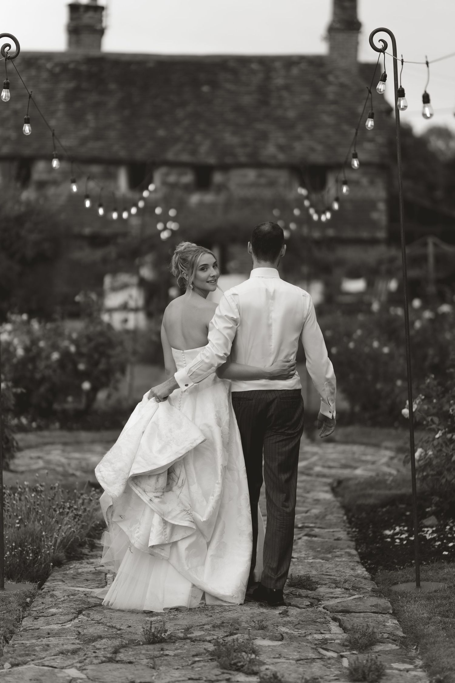 A romantic black and white photo of a couple walking along a garden path with twinkling lights above.