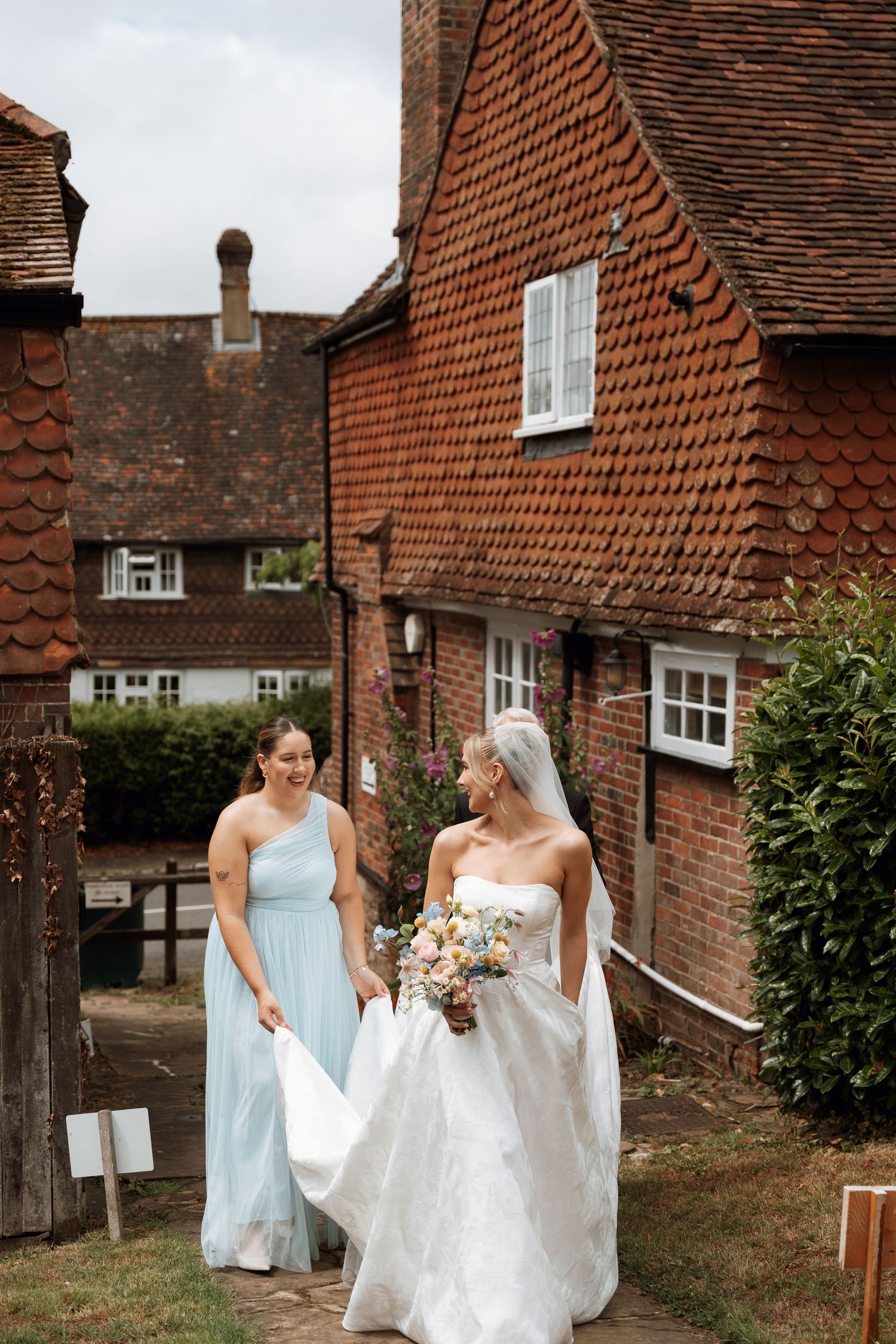 Bridesmaids in light blue dresses assist with a wedding gown outside traditional brick buildings.