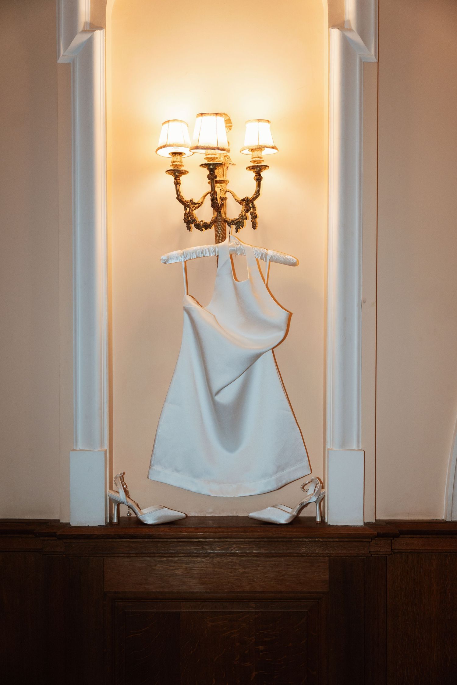 A white wedding dress hangs elegantly beneath an ornate wall sconce with glowing lamp shades in a warm-lit setting.