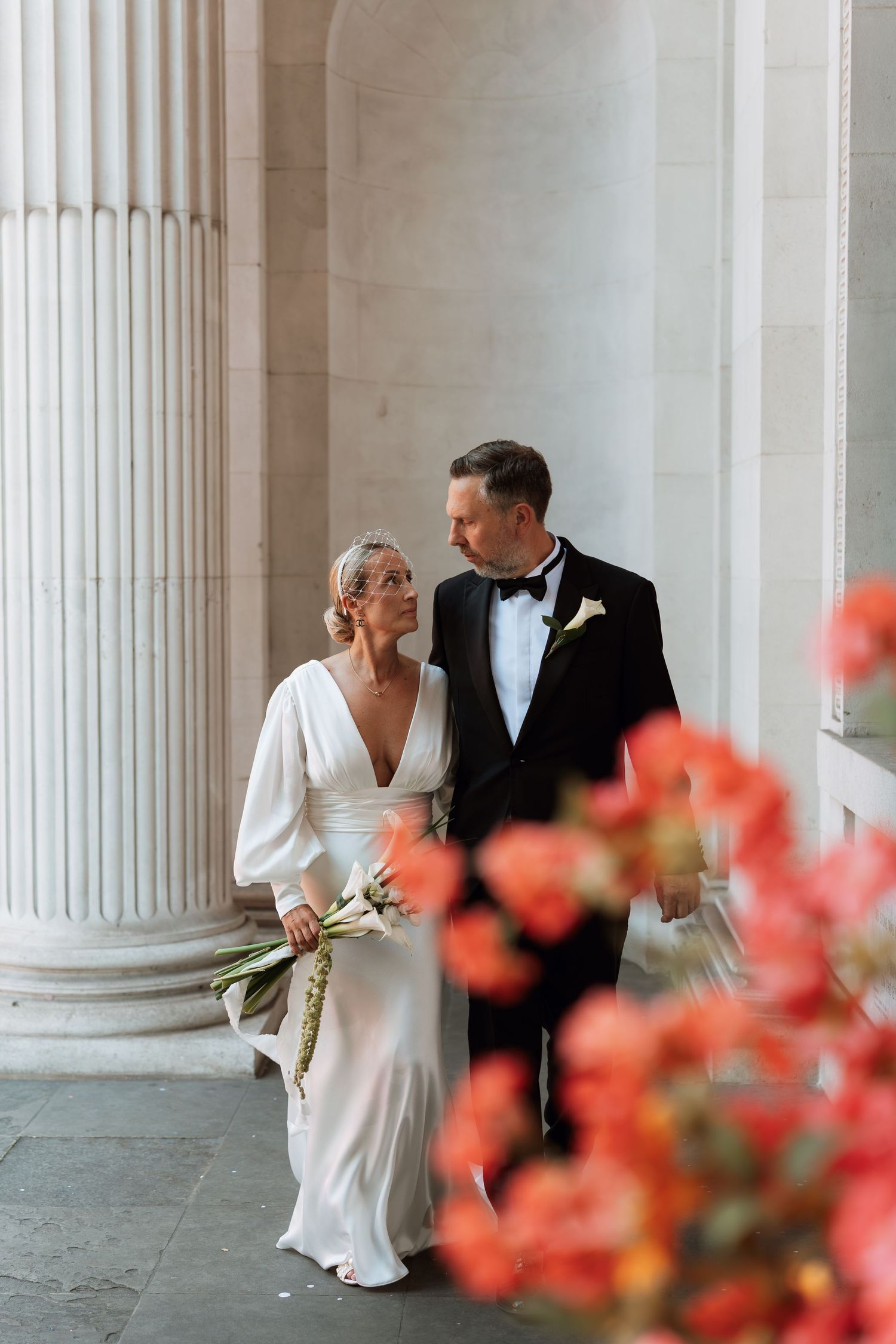Elegant wedding couple posing between classic white columns with coral flowers in foreground.