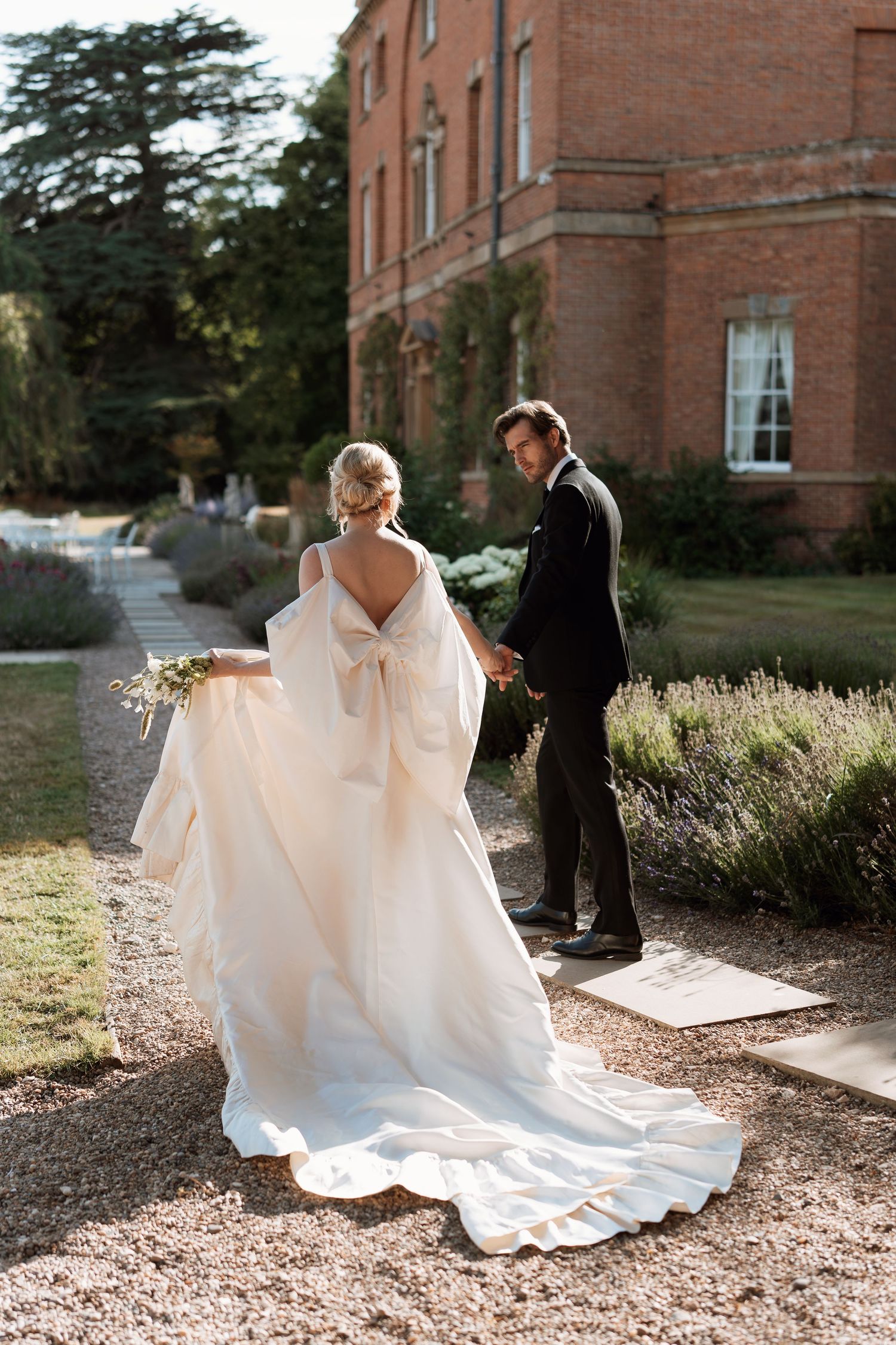 A wedding couple walks along a brick building as sunlight catches the flowing white wedding dress train.