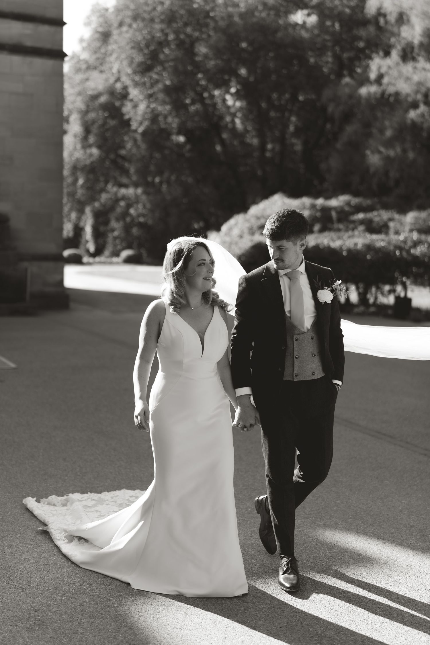 Black and white photo of newlyweds walking together on a sunlit pathway.