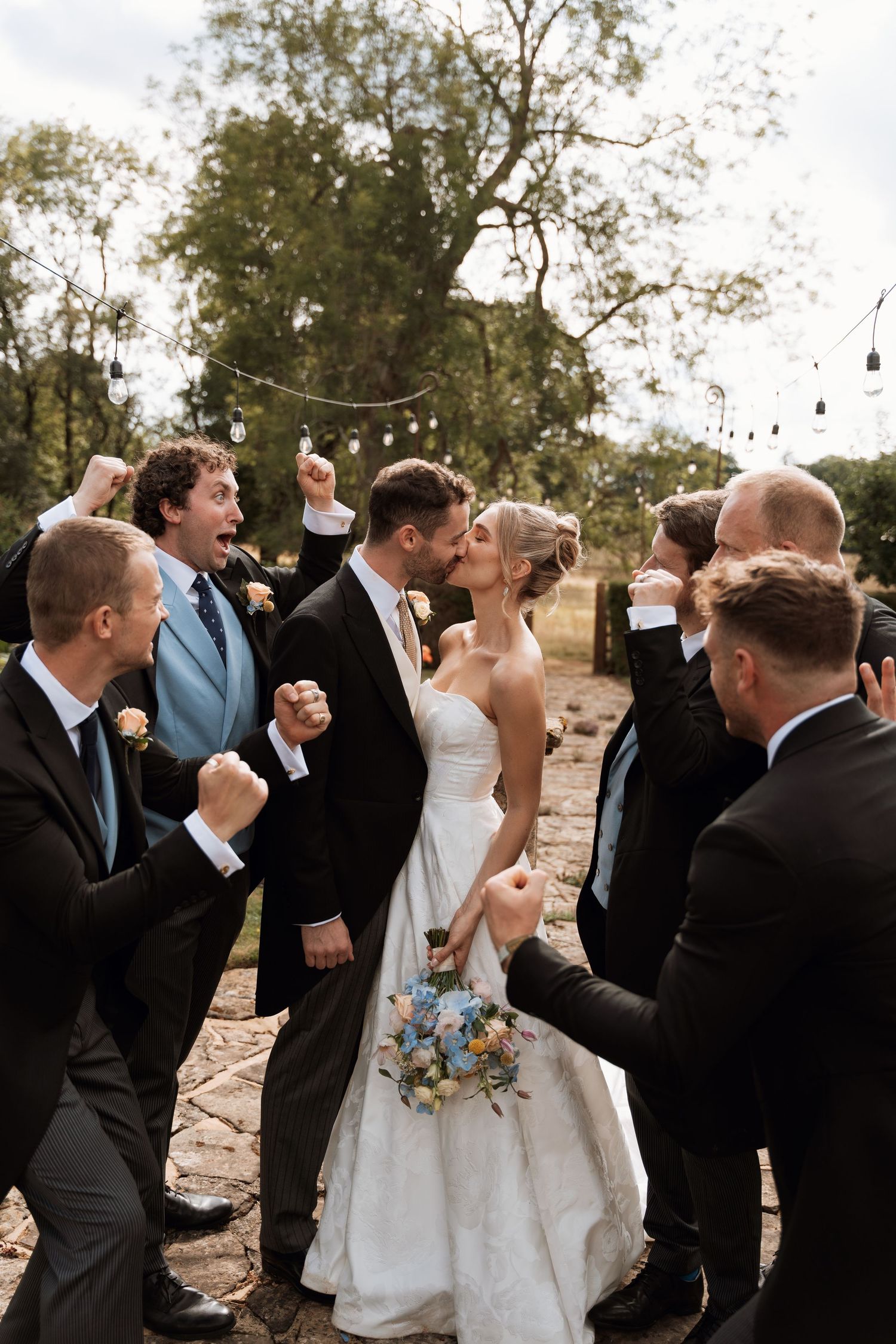 Wedding party celebrates with enthusiasm as the newlyweds share a kiss under string lights outdoors.