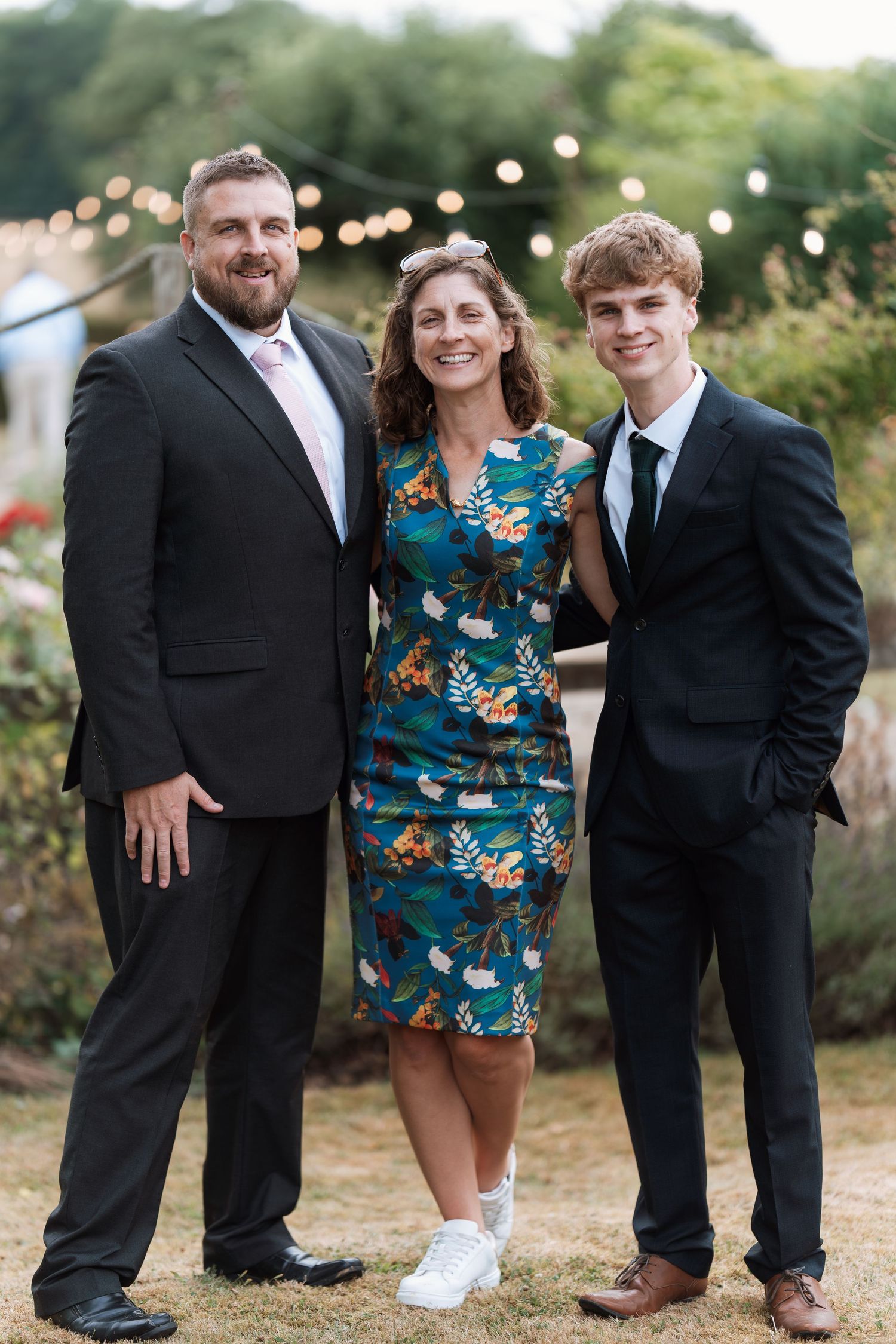 Three people in formal attire posing together at an outdoor event with string lights in the background.