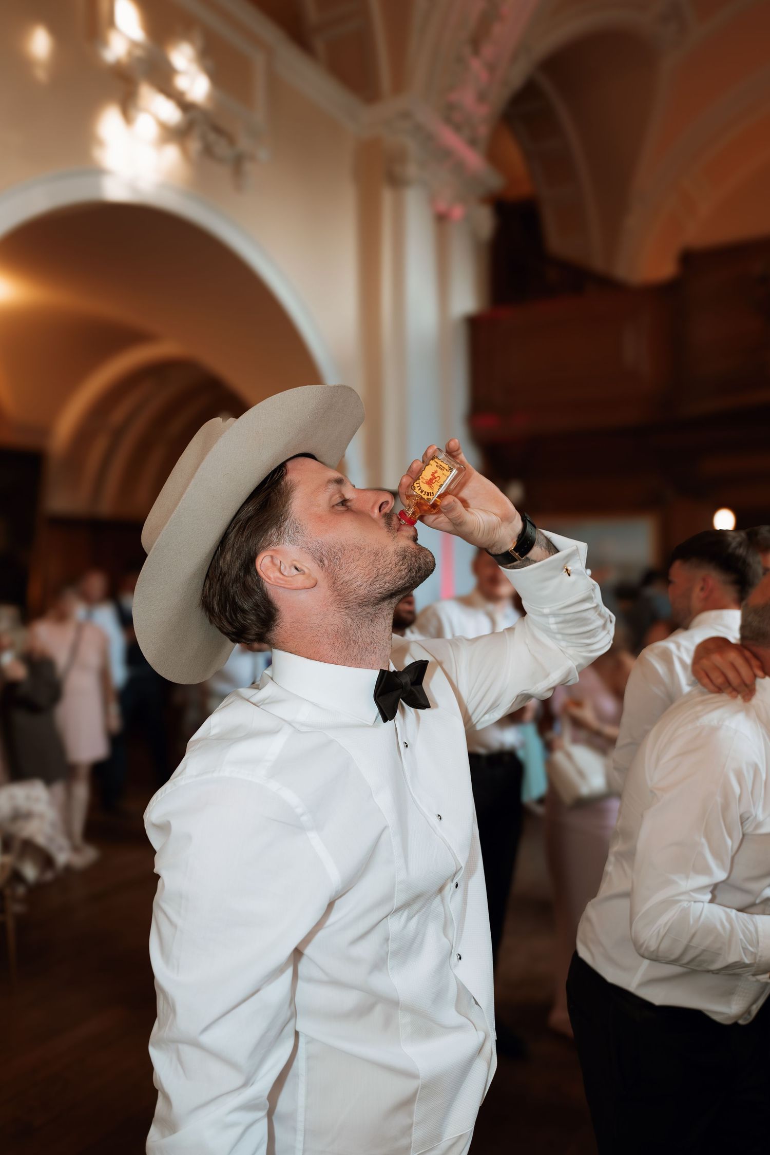 A guest in a white dress shirt and cowboy hat takes a drink at an elegant indoor wedding reception venue.