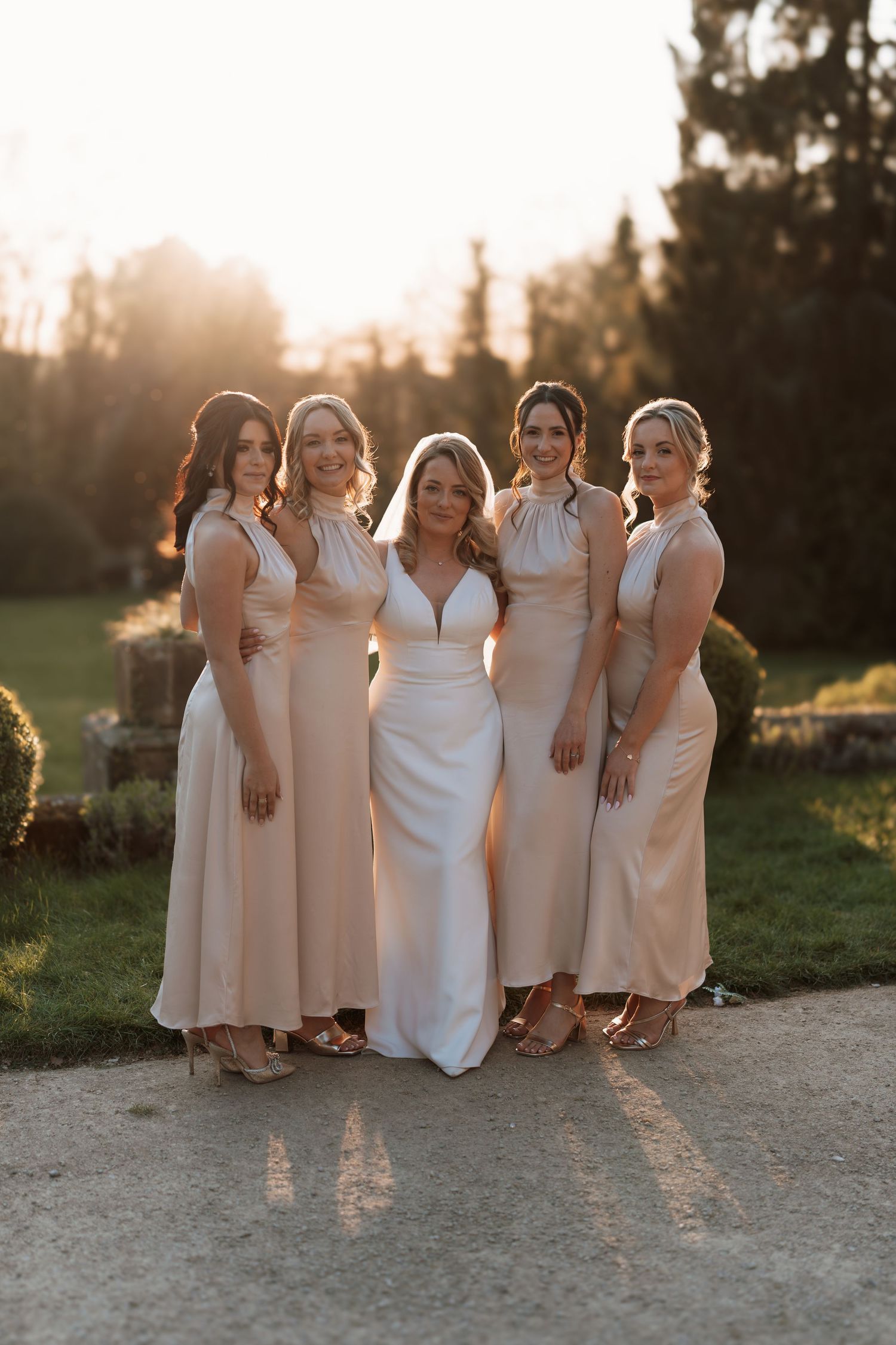 Bridesmaids in champagne-colored dresses pose with the bride in a white gown during golden hour.