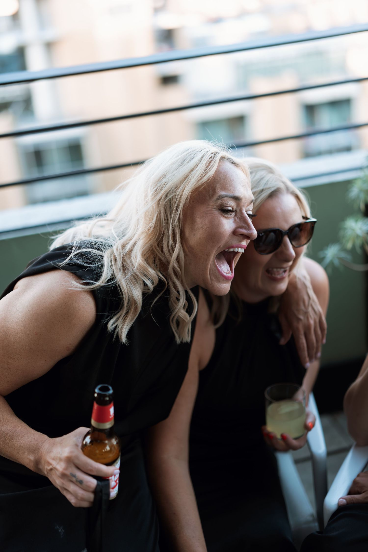 Two friends laughing and enjoying drinks on a balcony during sunset.