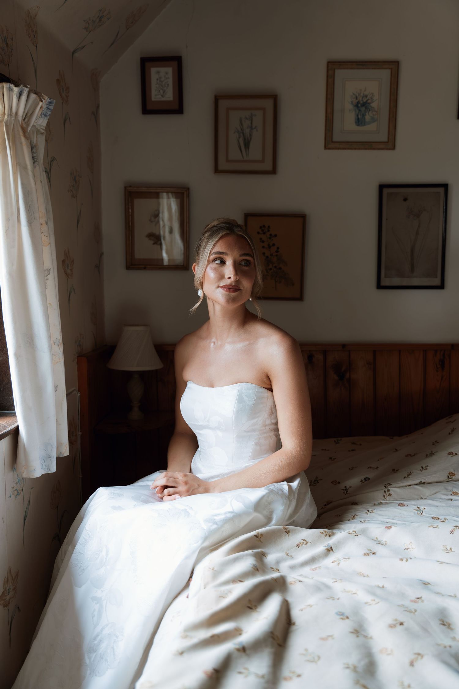 A classic bridal preparation moment captured in a rustic bedroom with vintage botanical prints on the wall.
