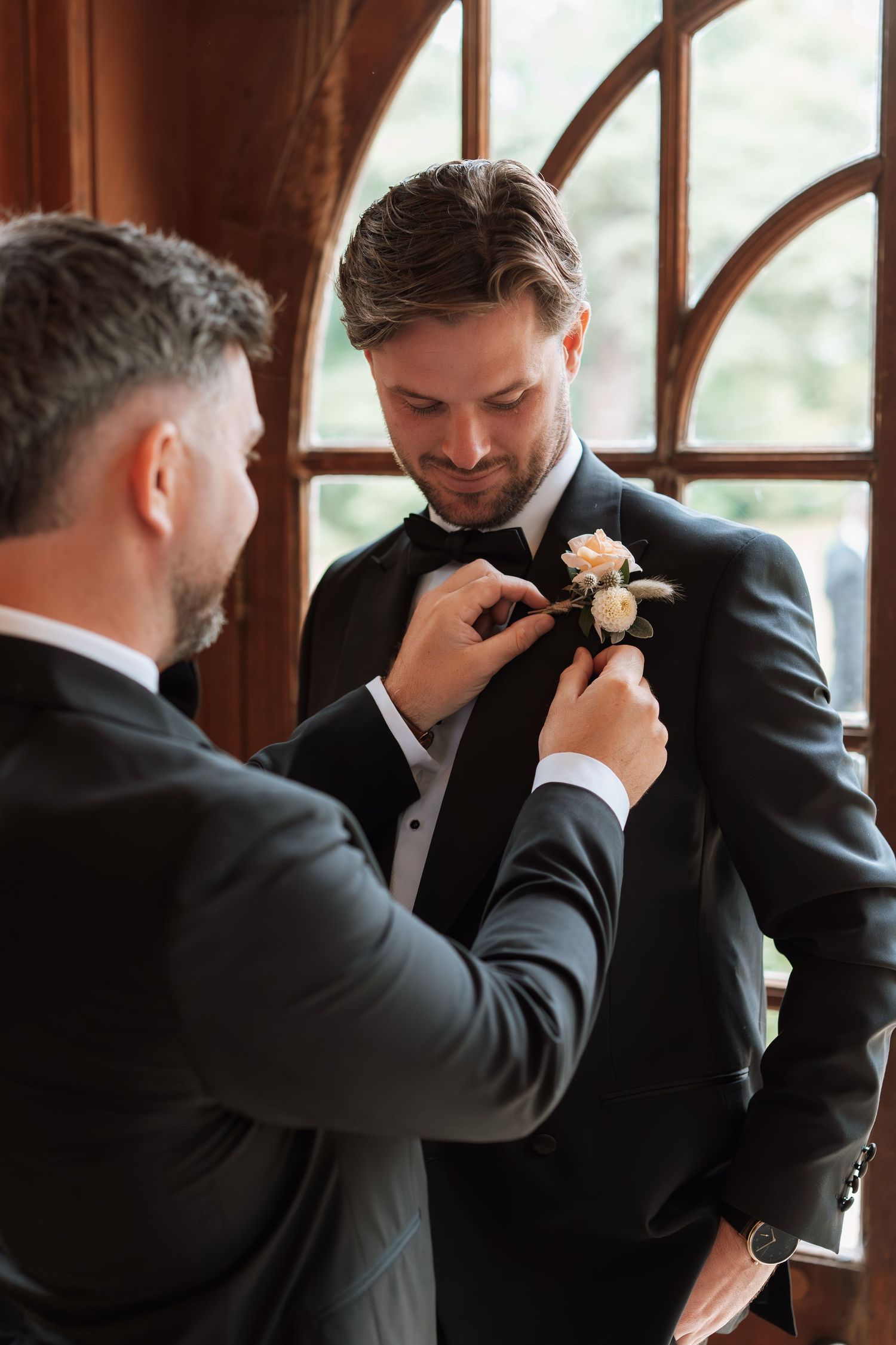 A groomsman helps adjust a boutonniere on another man's suit jacket lapel in front of an elegant arched window.