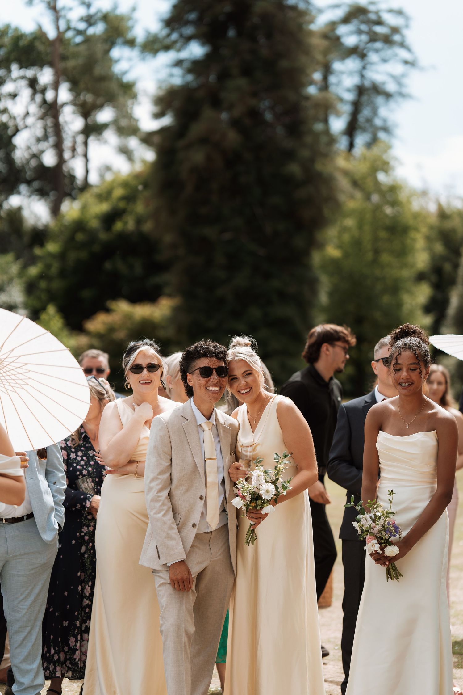 Wedding party in neutral cream and white attire poses in a line with parasols during an outdoor summer celebration.