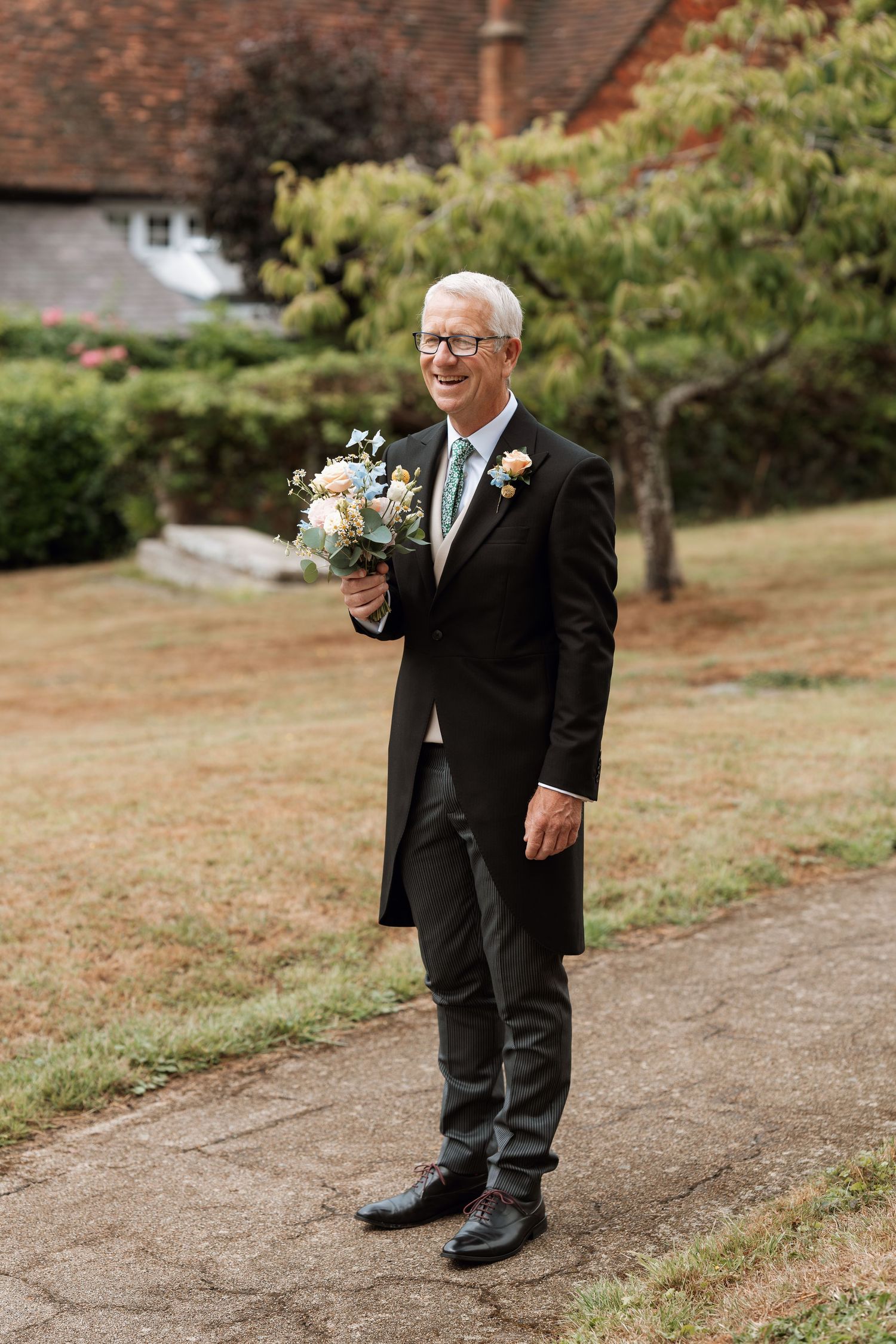A smiling member of the wedding party wearing a black suit holds a bouquet while standing outdoors on a stone path.