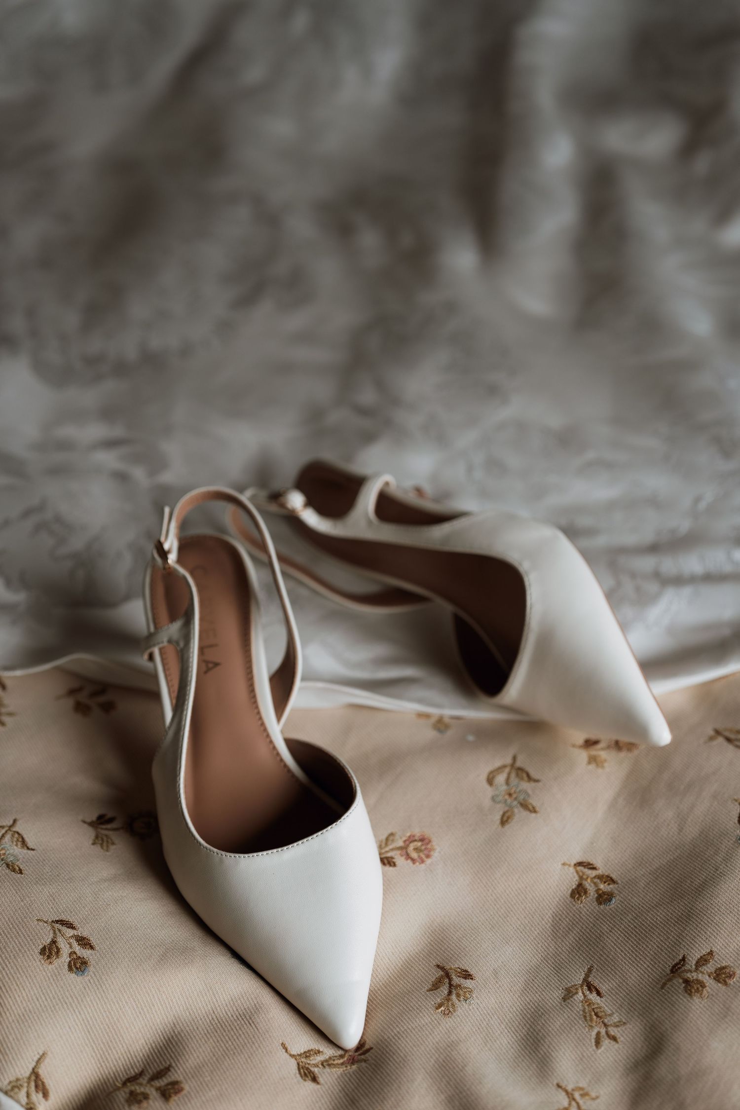 Elegant white pointed-toe slingback heels resting on a floral patterned fabric.