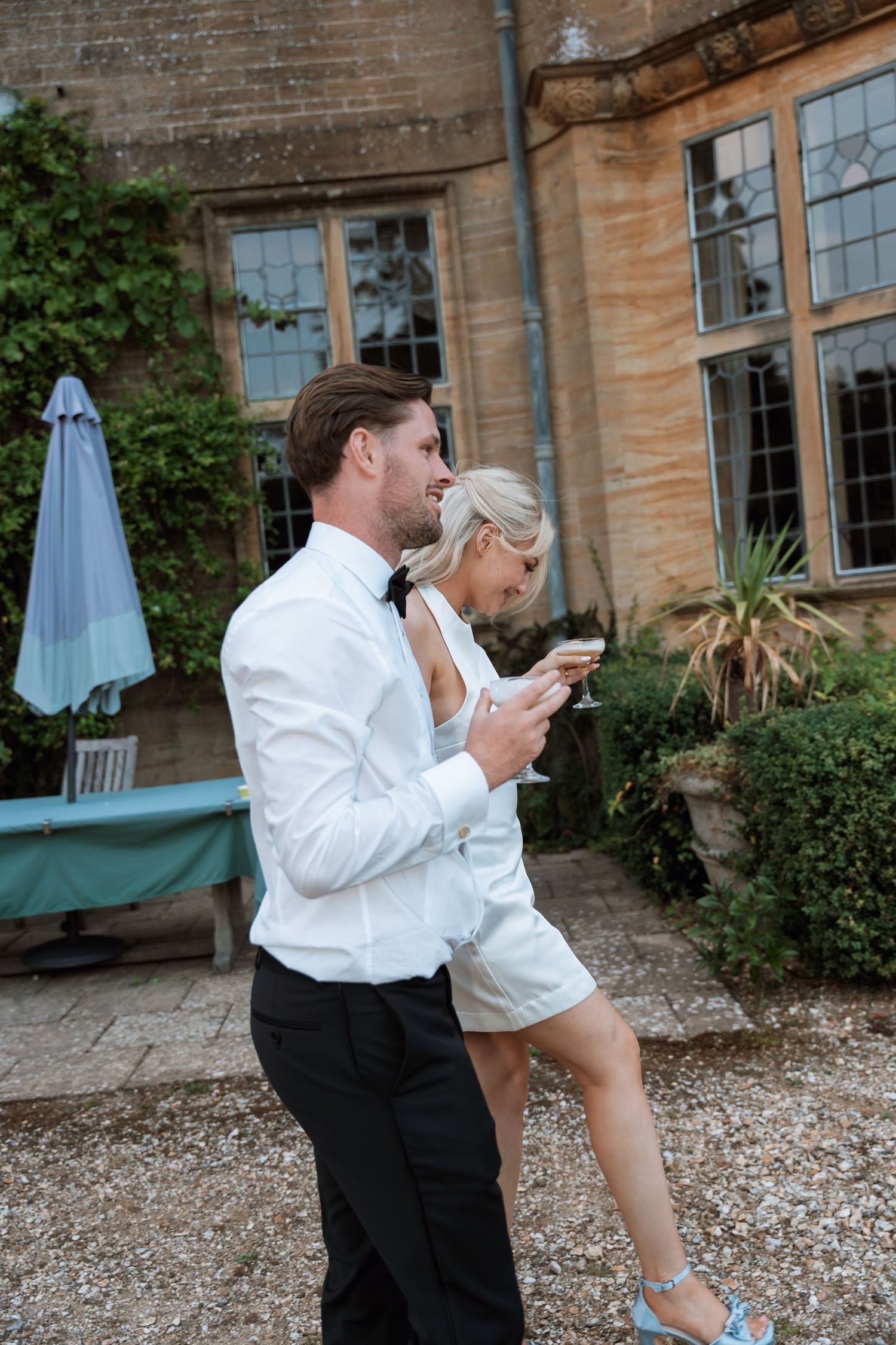 Two people in white outfits check their phones while standing on a gravel path outside a historic stone building.