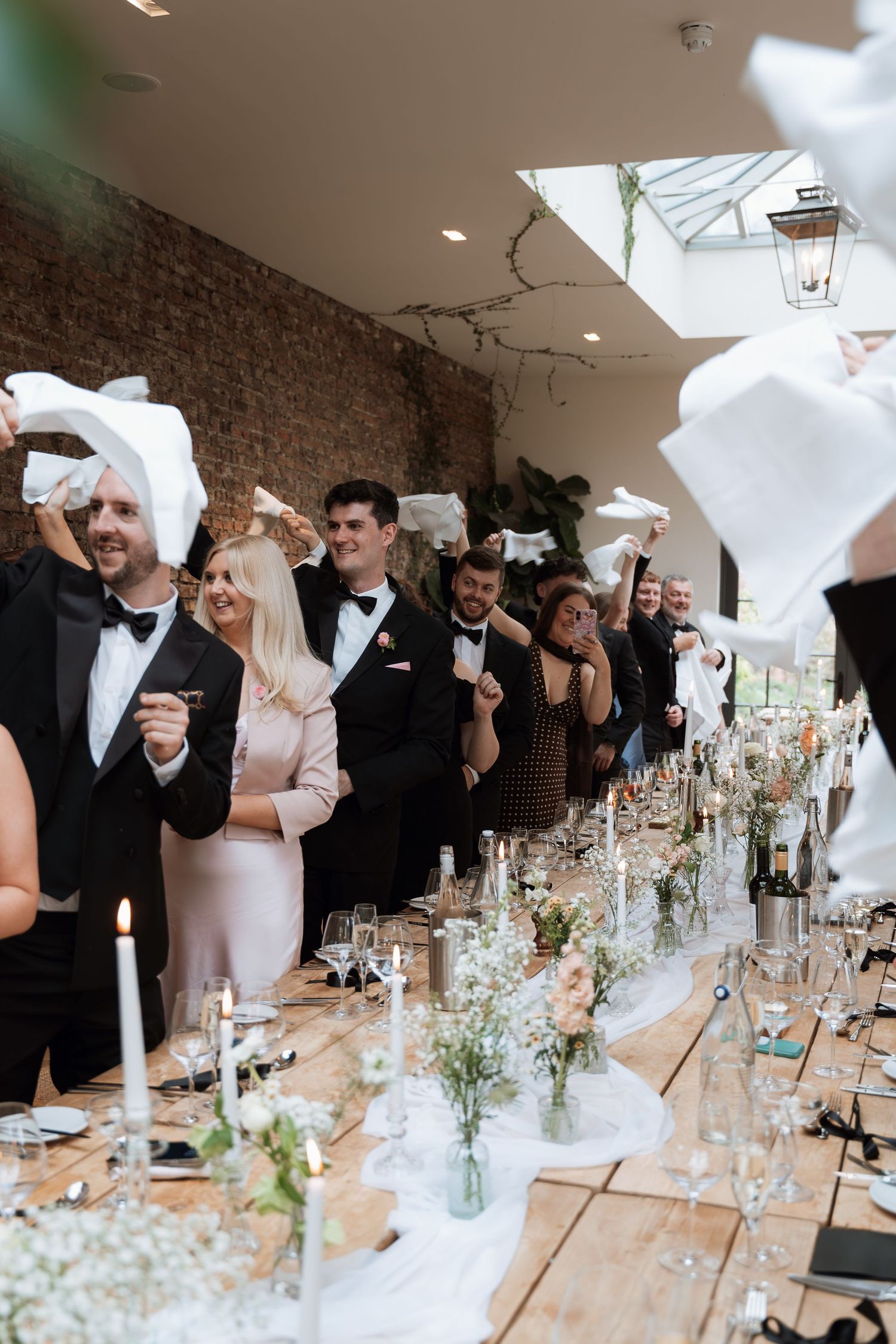 Festive wedding reception with guests raising glasses in toast along rustic wooden tables with floral centerpieces.