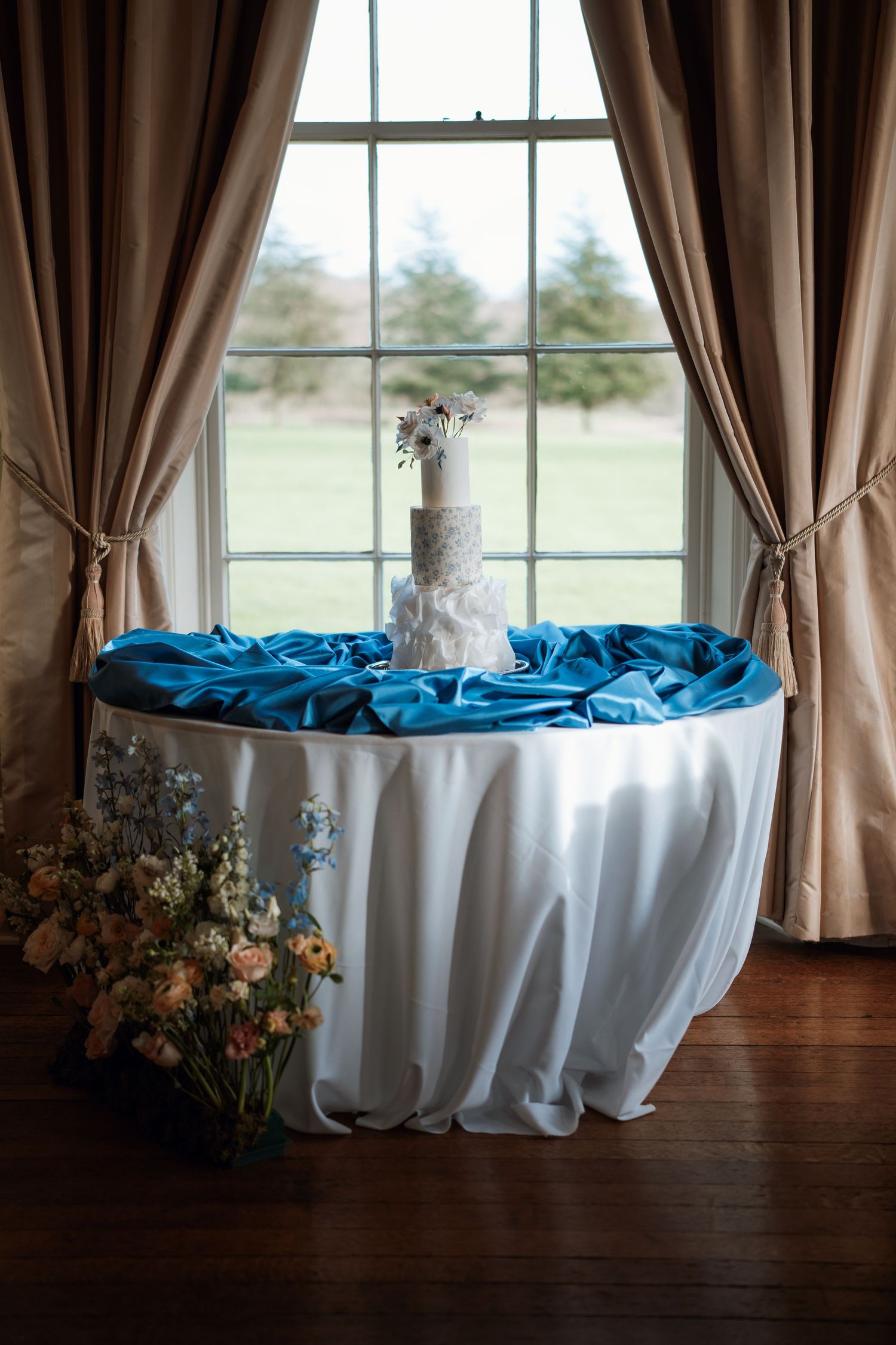 Elegant wedding table with white tablecloth, blue accents, and floral centerpiece by window.