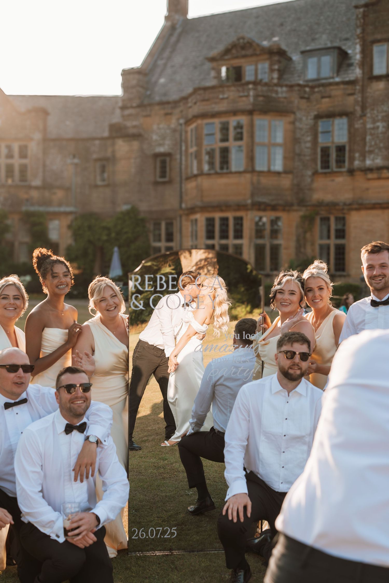 Wedding party celebrates on sunny lawn in front of grand historic stone mansion at golden hour.