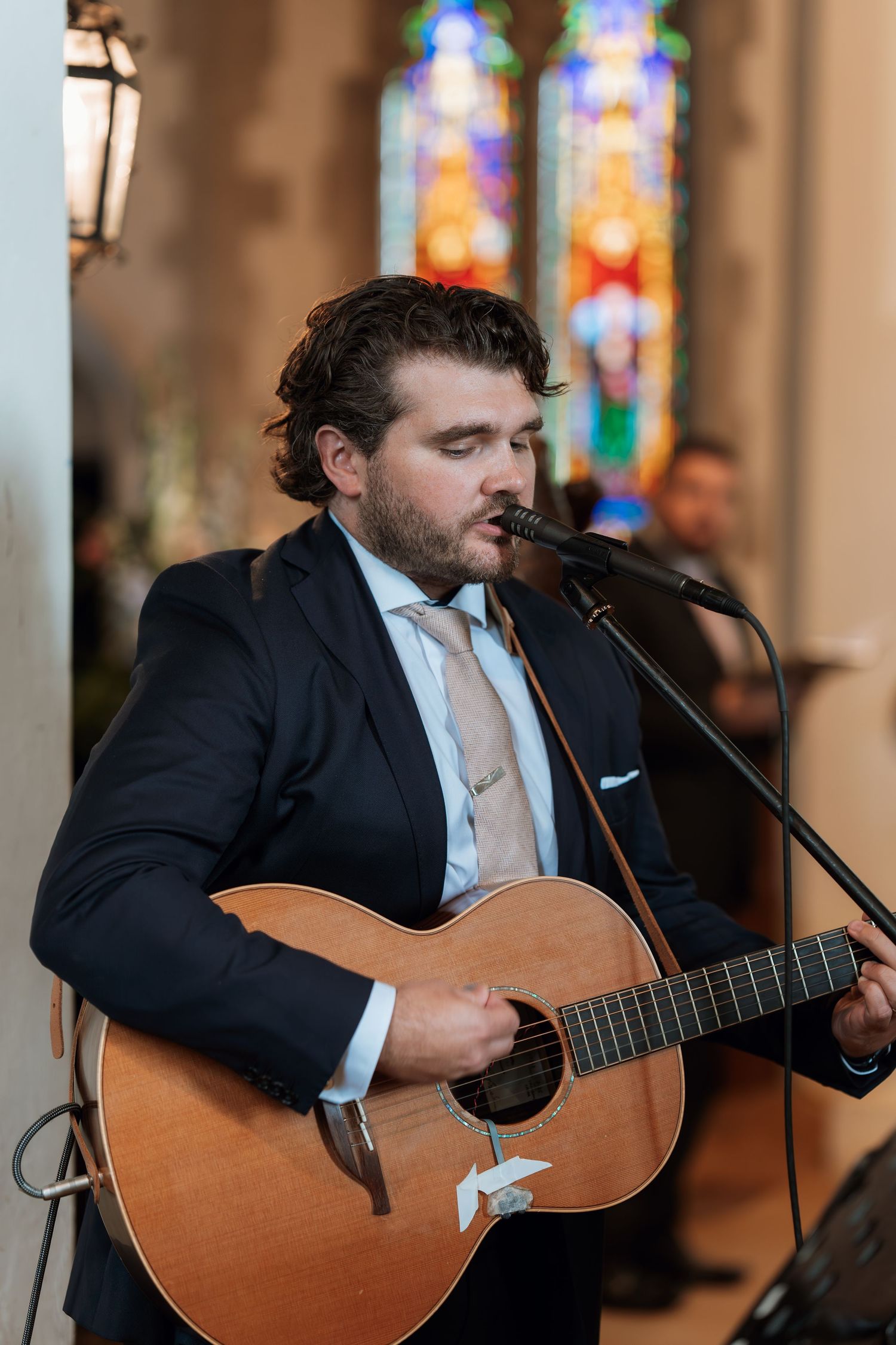 Musician in formal attire performs with acoustic guitar in front of stained glass windows.