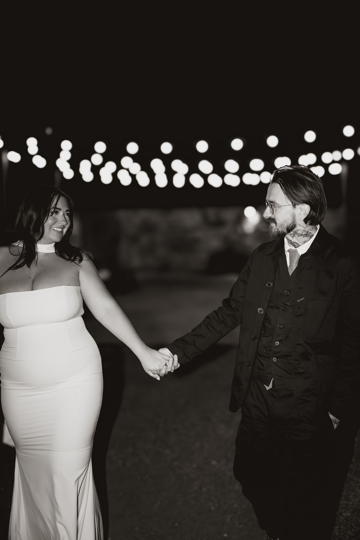 Black and white wedding photo showing figures dancing under string lights at an outdoor evening reception.