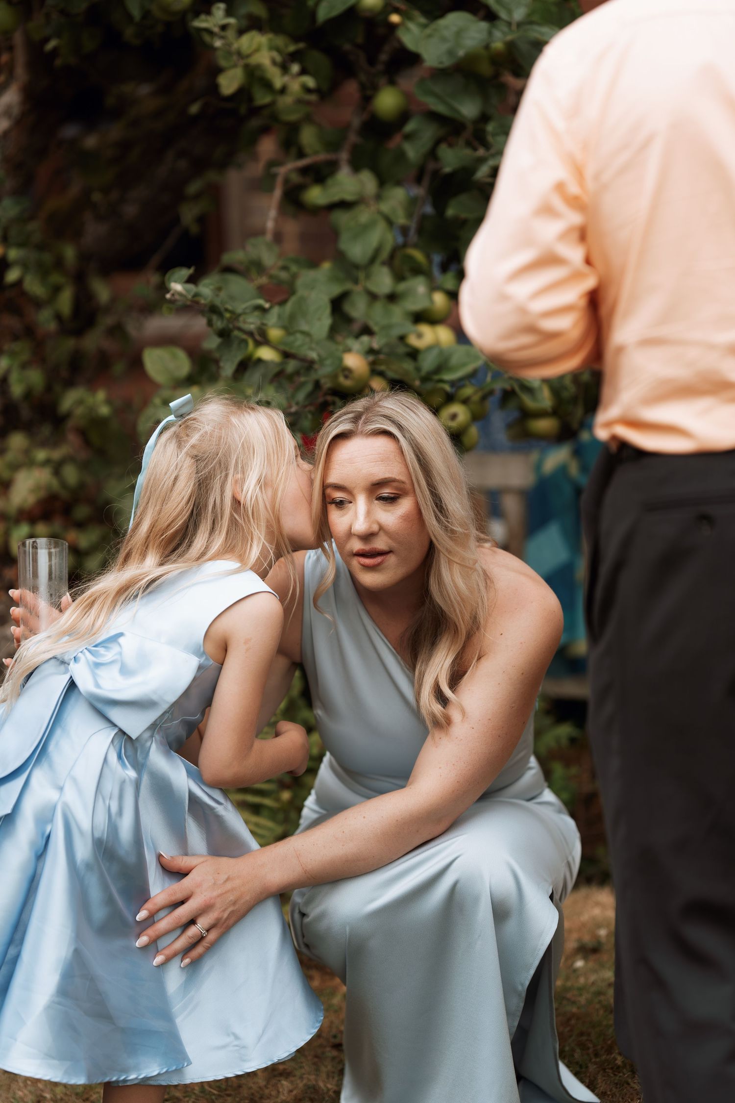 A tender moment between an adult and child in light blue dress sharing a secret in an outdoor garden setting.