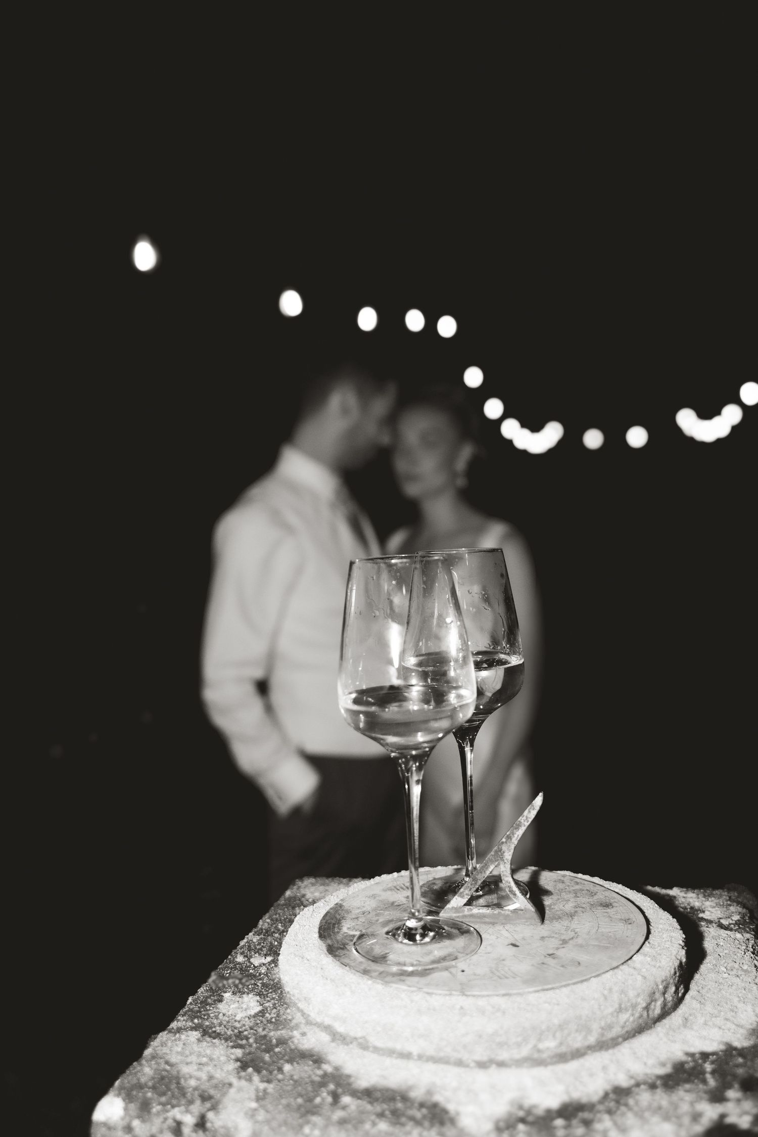 Two wine glasses on a stone surface with string lights and a romantic silhouette in the background.
