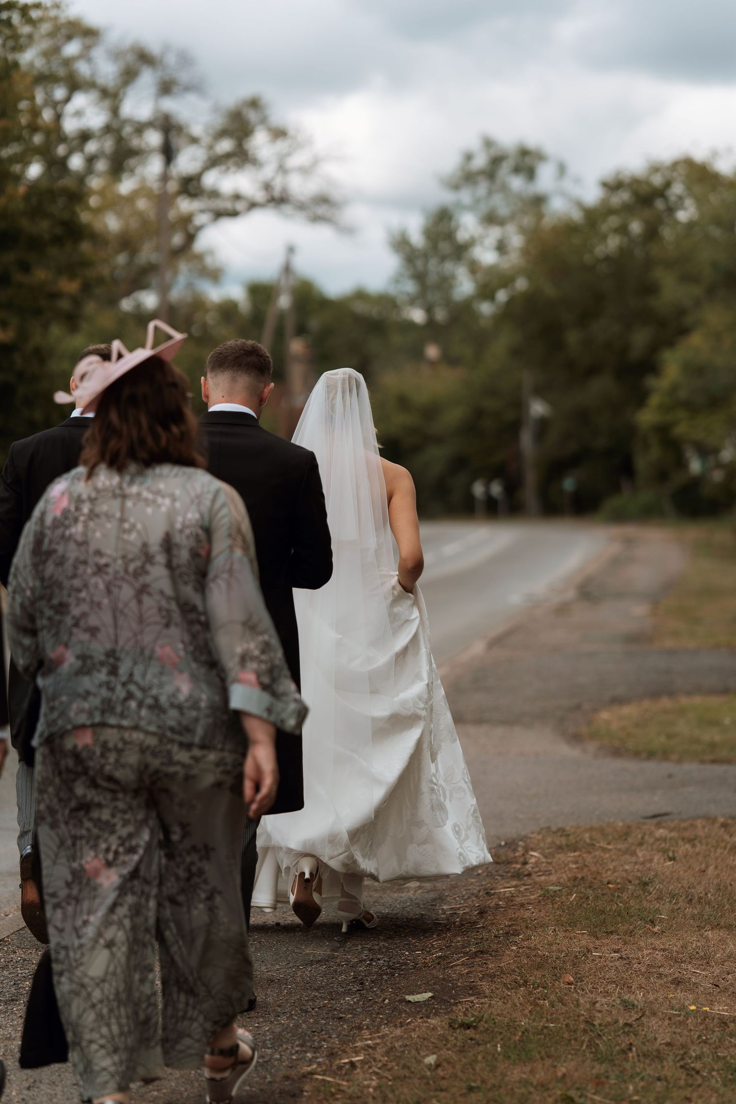Wedding party walks along a countryside path under cloudy skies with flowing white gown trailing behind.