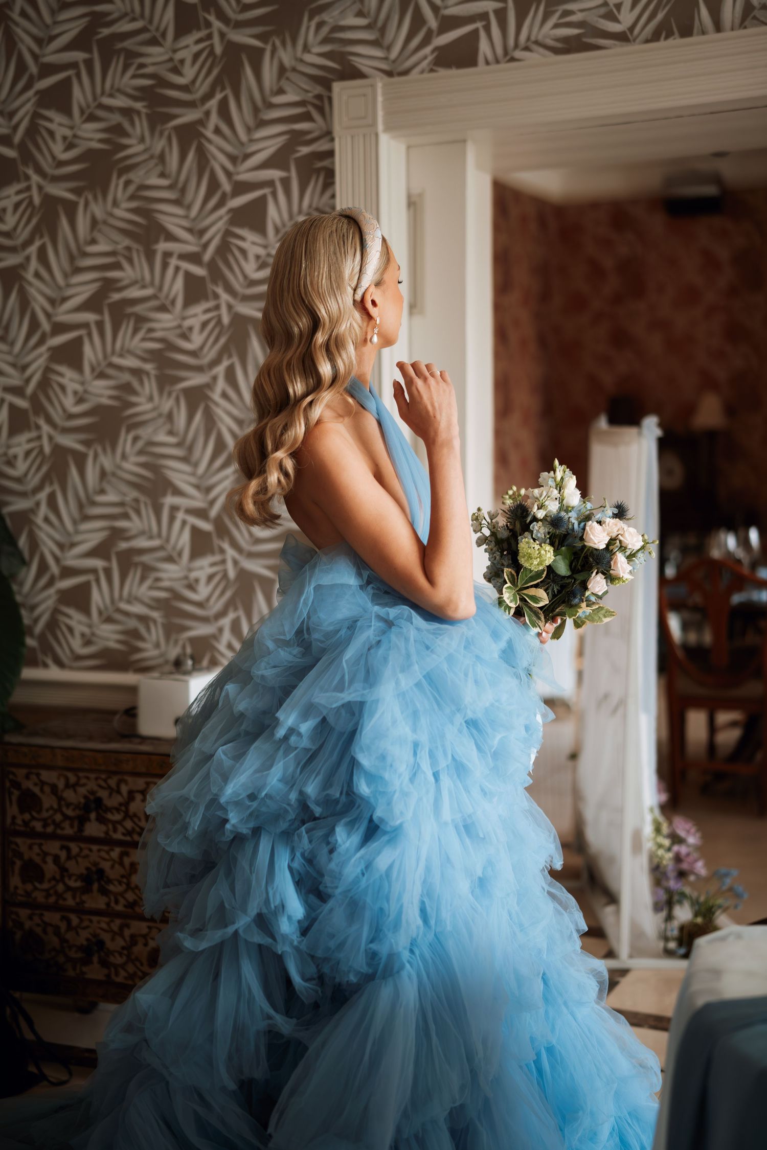 A stunning light blue tulle ballgown with ruffled layers sits elegantly against floral wallpaper.