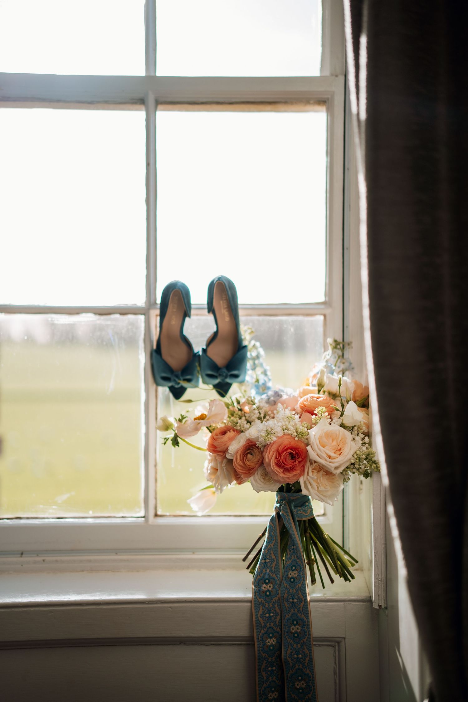 Elegant blue wedding shoes and a peachy pink bouquet rest on a windowsill.