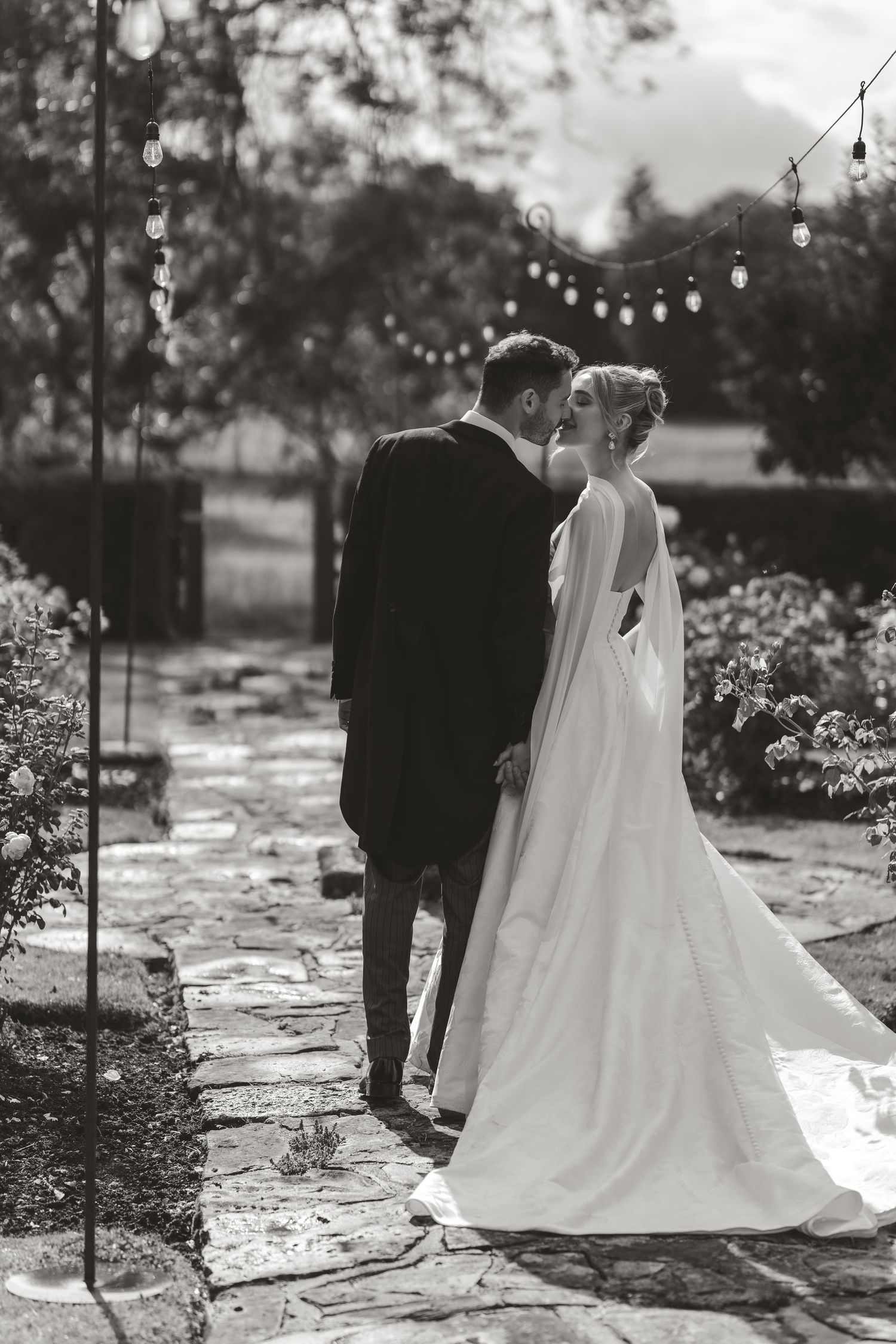 A black and white photo of newlyweds walking along a stone path under romantic string lights.