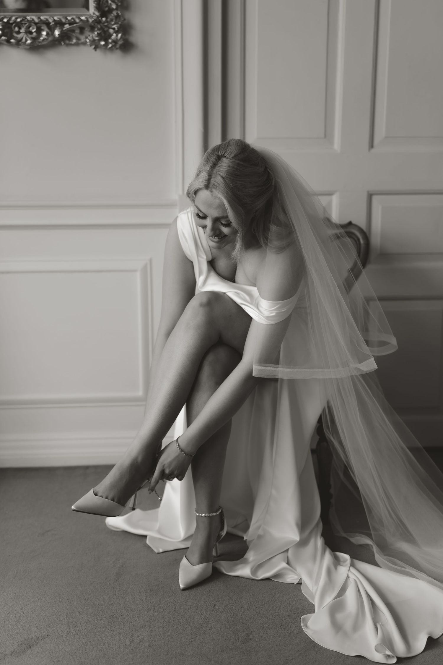 Black and white photo of a bride in a wedding dress and veil fastening her shoe while seated.