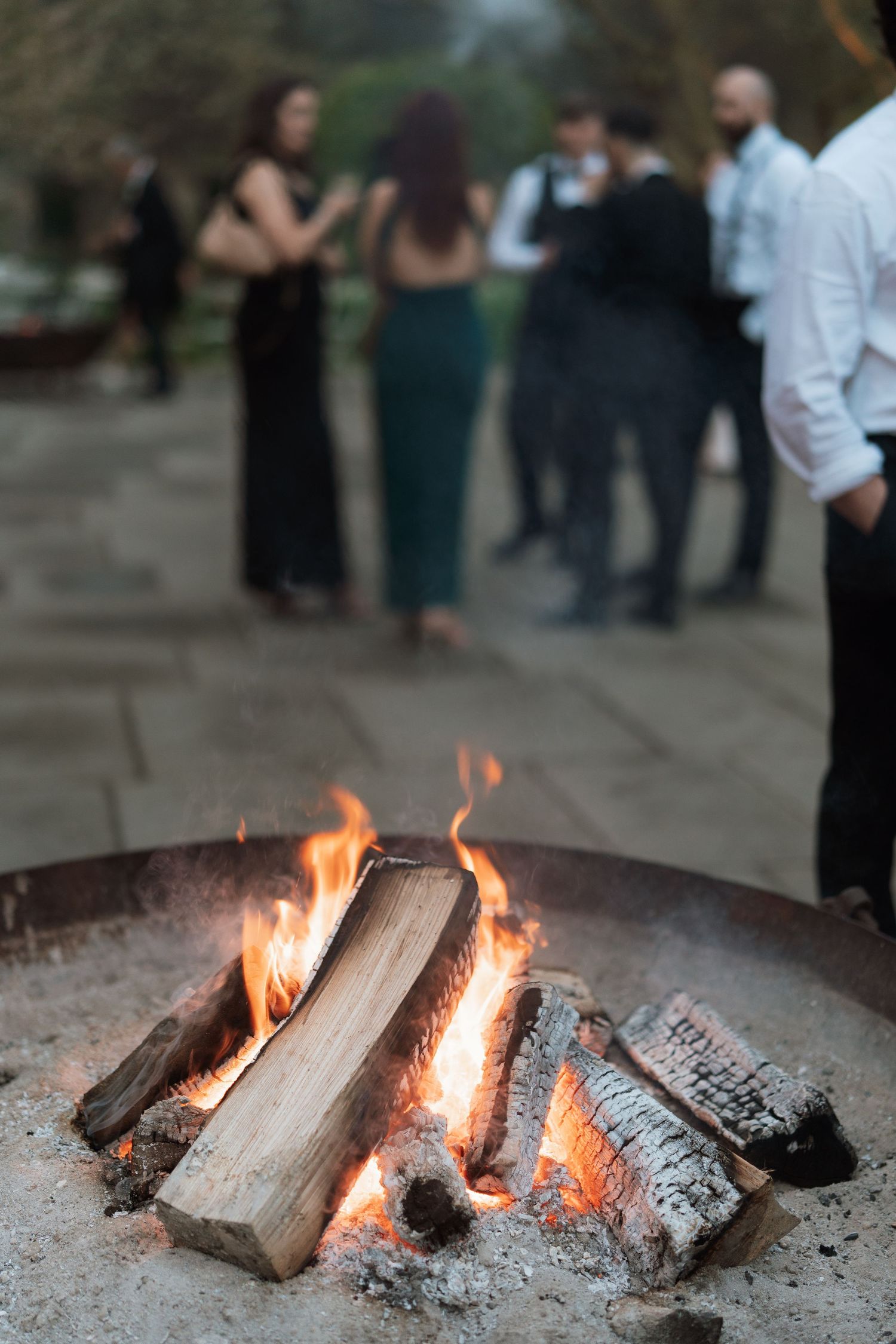 Burning logs in outdoor fire pit with party guests mingling in background.