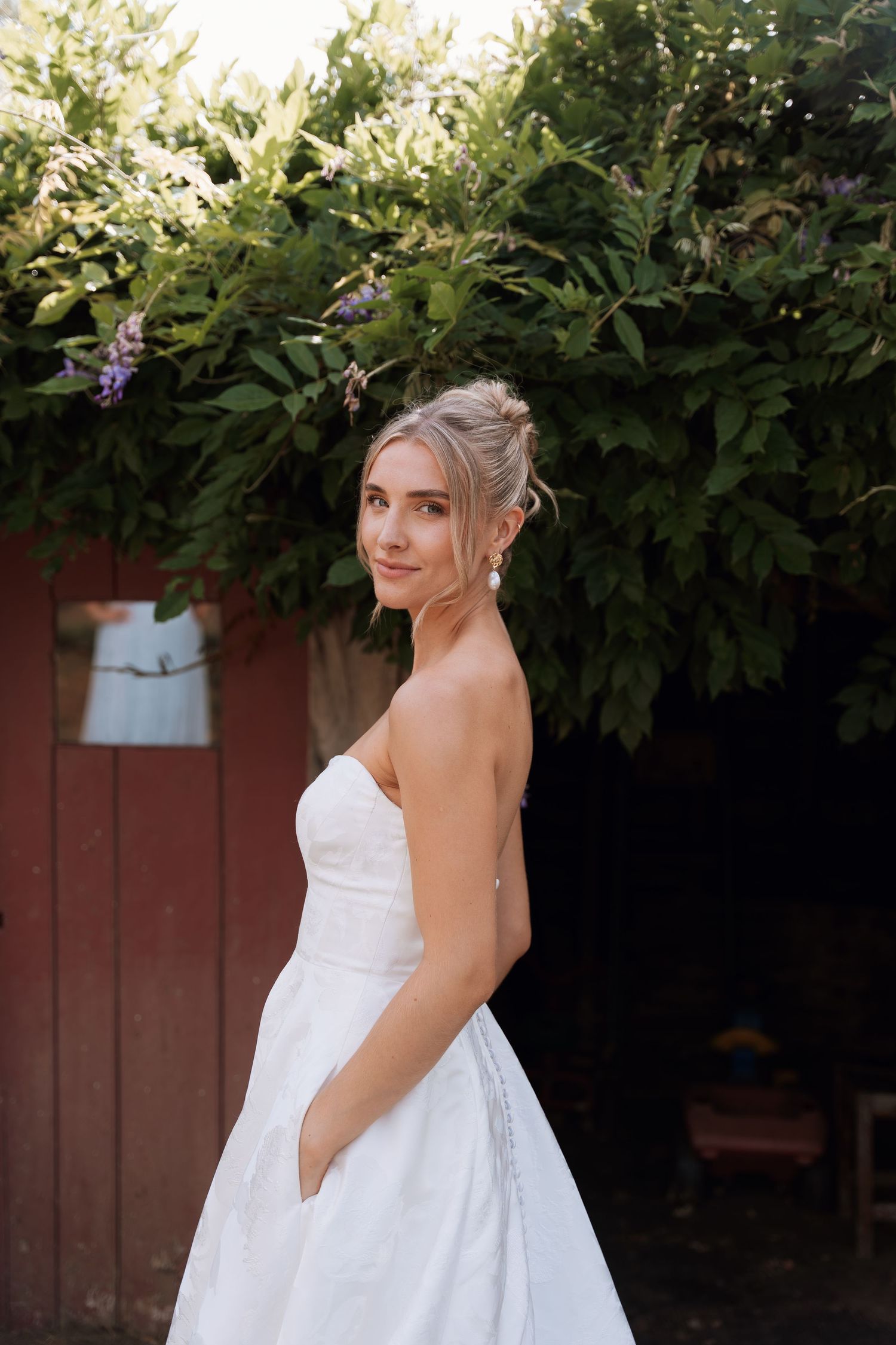 Elegant white wedding dress photographed against lush green foliage and red fence backdrop.
